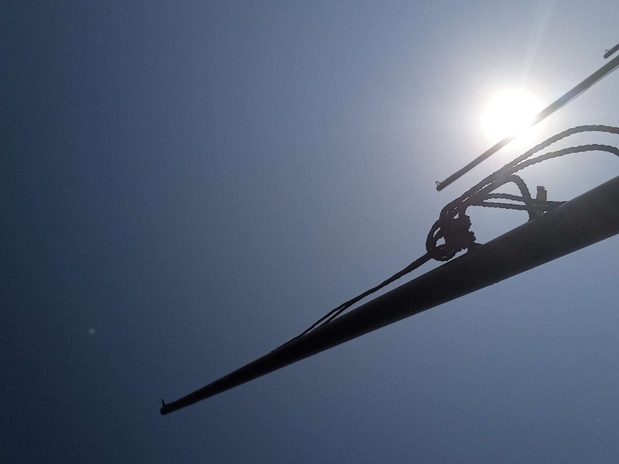 The image shows a silhouette of a flagpole against a bright, clear blue sky. The sun is partially obscured by the flagpole and appears as a bright orb with rays emanating outwards. A thick rope is attached to the flagpole, with knots visible. There is a faint, out-of-focus circular shape in the upper left portion of the sky, possibly a distant object or atmospheric phenomenon. The perspective is from below, looking upwards. The scene is likely outdoors. There are no people or animals visible. The image is taken in Bububu, Tanzania.