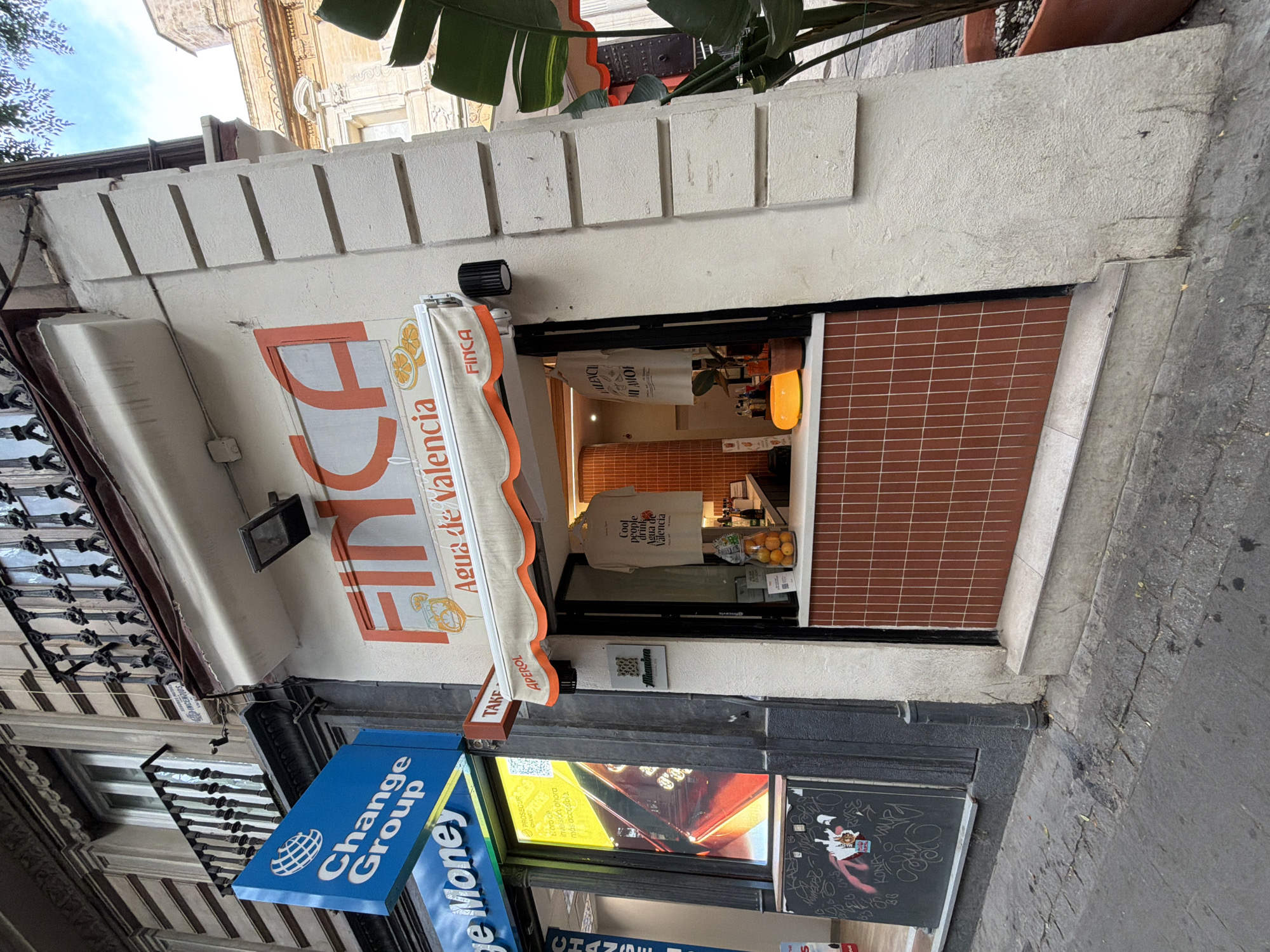 The image shows a storefront with a sign that reads "FINCA Agua de Valencia". The storefront appears to be a cafe or juice bar, with a display of t-shirts and fruit inside. To the left of the storefront is another business with a sign that says "Change Group". The scene is located on a city street with cobblestone pavement.