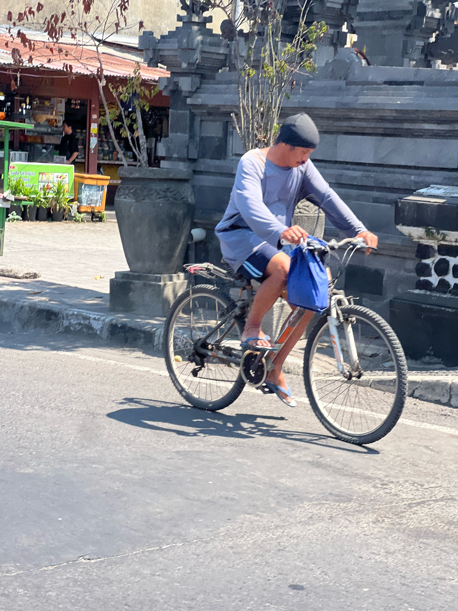 A man wearing a blue long-sleeved shirt and dark shorts rides a bicycle with a blue bag hanging from the handlebars. He is wearing a dark beanie and flip-flops. The bicycle has a white frame with orange accents. The man is on a paved road in Denpasar, Indonesia, with a building and a large stone planter in the background. The building appears to be a shop with awnings, and the planter contains a small tree. The scene is brightly lit by sunlight, casting shadows on the road.