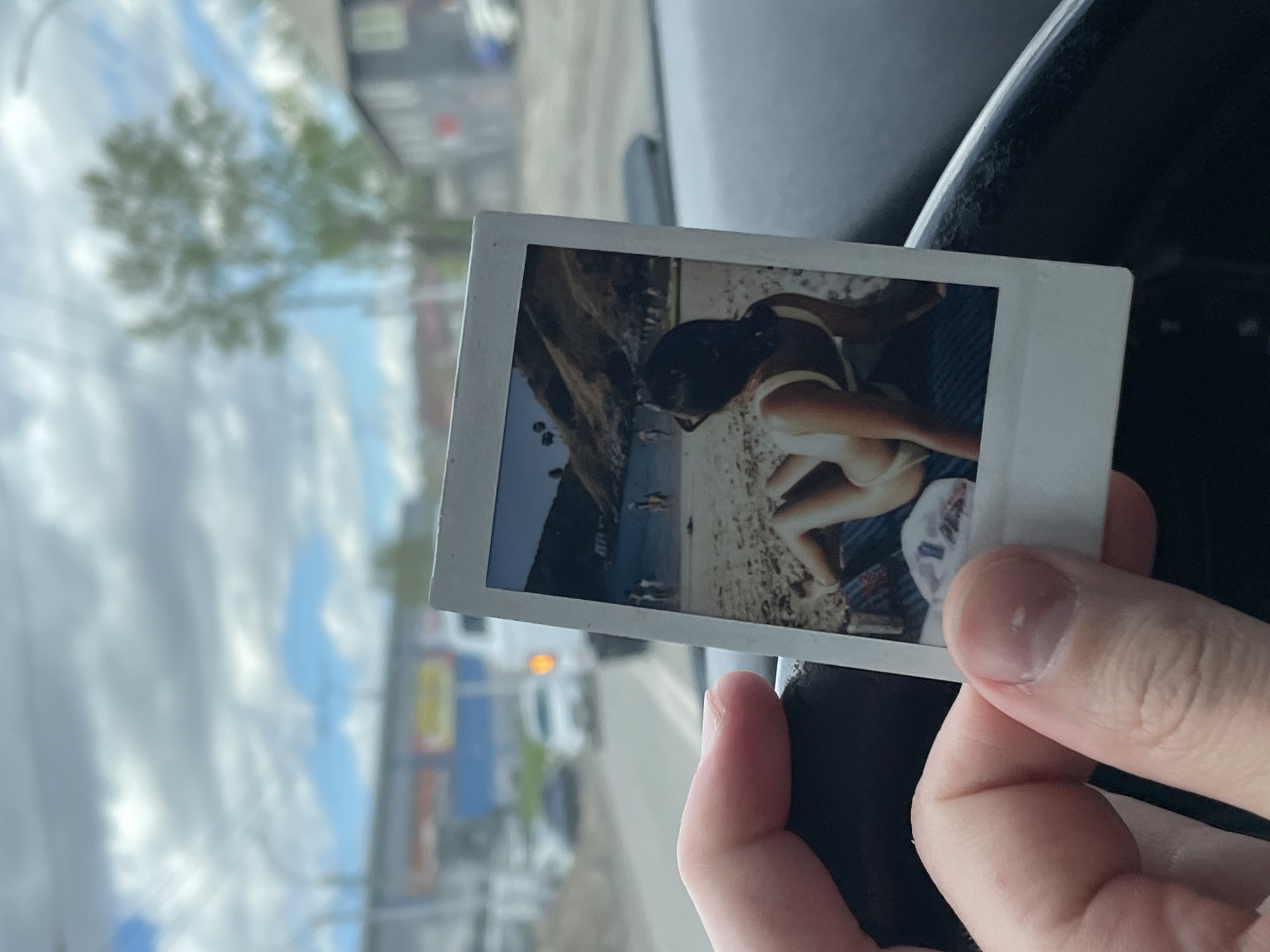 A hand holds a Polaroid photograph displaying a person in a bathing suit on a beach with an ocean backdrop. The Polaroid is held in a car interior, visible near the steering wheel. Outside the car window, a blurred urban landscape is visible, including trees, buildings, a blue and yellow sign, and other vehicles. The location is undetermined.