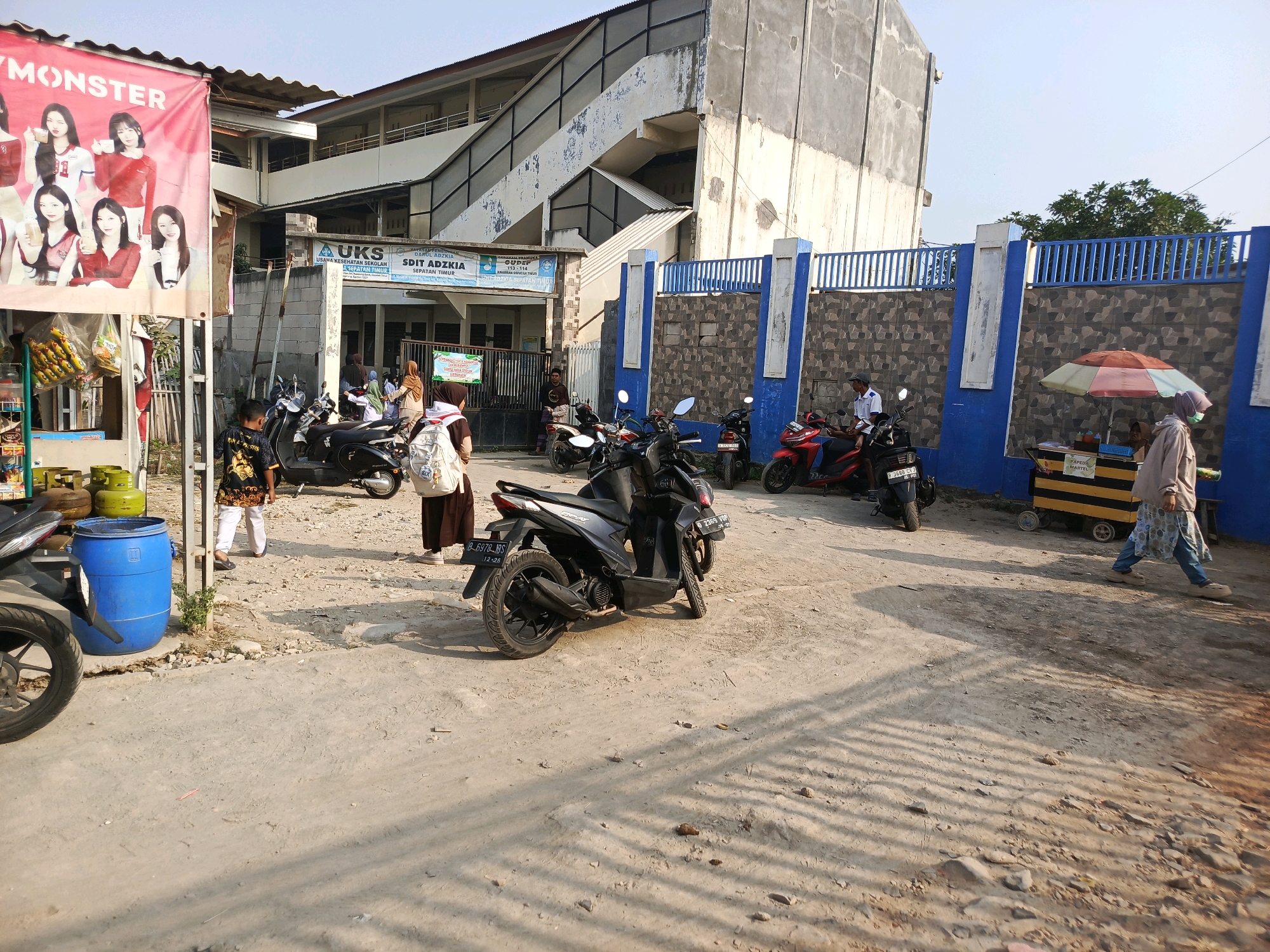 The image depicts a scene outside a school in Jati Mulya, Indonesia. In the foreground, a dirt and gravel area is visible with several parked motorcycles. A blue plastic barrel and propane tanks are also present on the left. A boy wearing a patterned shirt and white pants walks across the area.
To the right of the barrel, a group of people are seen standing. A young boy in a black shirt with a gold pattern walks away from the camera. Two young women, one wearing a headscarf and a long brown skirt with a white backpack, and another in a white headscarf and a brown abaya, stand together.
Further into the background, a building with multiple floors and an open-air staircase is visible. A blue fence runs along the front of the school grounds. Several more motorcycles are parked in front of the fence.
On the right side of the image, a food cart with a colorful umbrella is set up. A person wearing a light brown hooded jacket and blue jeans walks past the cart, and a person in a white shirt is seated on a red motorcycle.
A large banner with images of several young women is displayed on the left side of the frame, advertising "MONSTER." The overall impression is that of a busy school entrance or exit area with vendors and students present. The sky is clear and the sun appears to be shining, casting shadows on the ground.