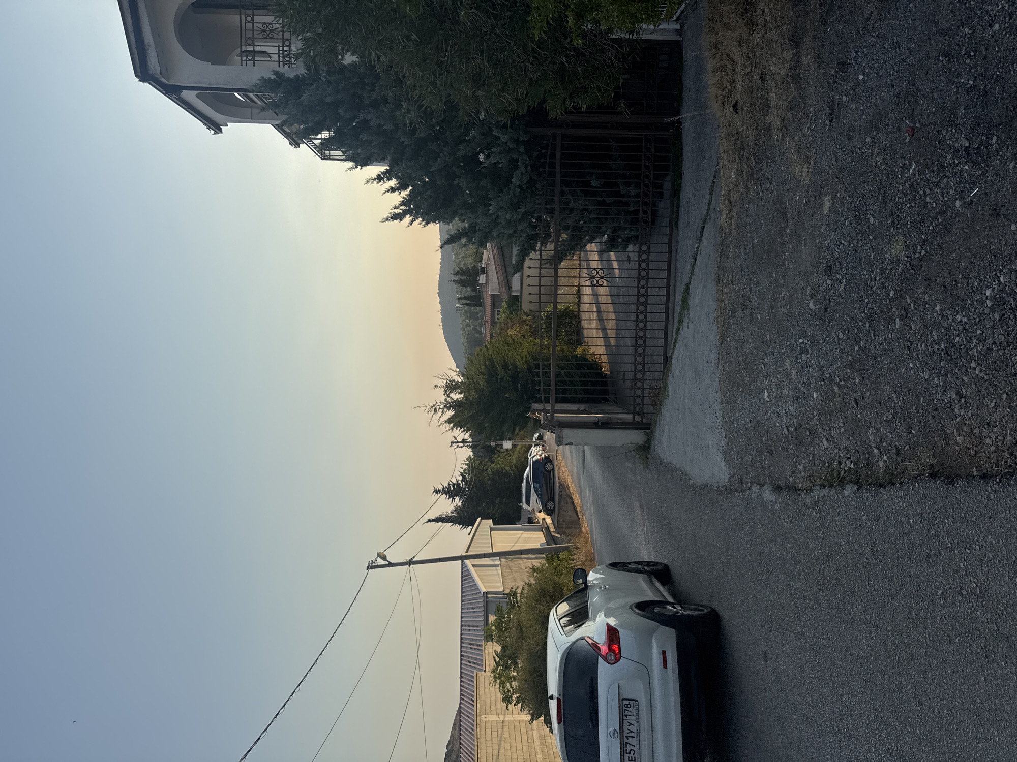 The image depicts a street scene in Sutomore, Montenegro, at what appears to be either sunrise or sunset, given the warm, soft lighting. The foreground is dominated by a white Nissan Juke car parked on the right side of a paved road, its rear end facing the viewer. The road curves gently to the left, leading into a residential area.
To the left of the road, a low, light-colored building with a corrugated roof is visible, partially obscured by some vegetation. Further down the street, a few other cars are parked, including a dark-colored hatchback. Electrical poles and overhead wires run along the left side of the road.
On the right side of the image, a dark, ornate metal gate stands closed, leading into a property. Behind the gate, tall, dark green cypress trees are prominent. Beyond the trees, a multi-story building with arched windows and balconies is visible, suggesting a villa or apartment complex. In the background, rolling hills are covered with vegetation and scattered houses. The sky is a clear, pale blue, with a hint of orange near the horizon, reinforcing the impression of early morning or late evening. The overall impression is one of a quiet, residential street in a Mediterranean coastal town.