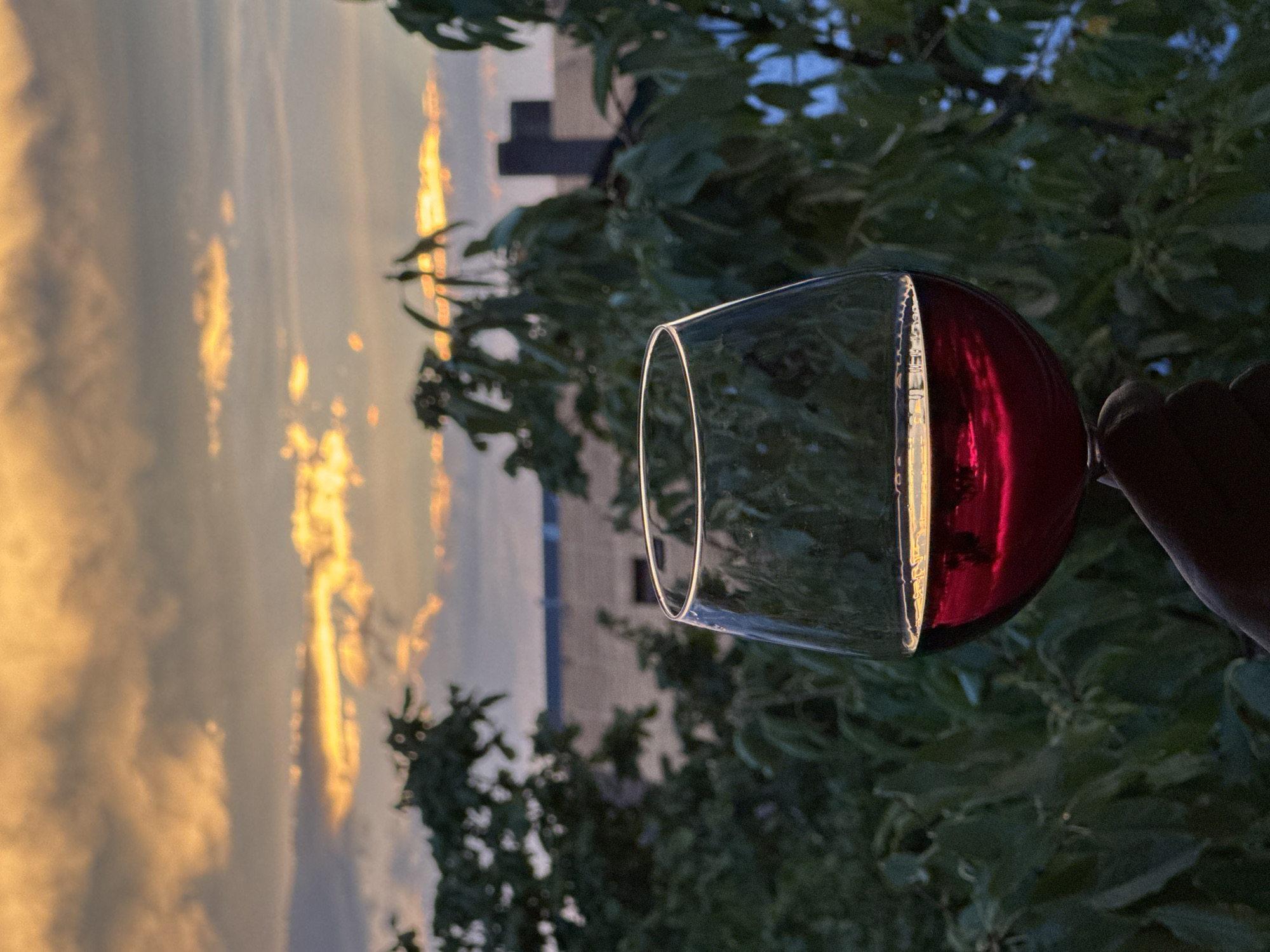 A hand holds a wine glass filled with red wine. The wine glass is in the foreground, and the background shows a blurred sunset with orange and yellow clouds, silhouetted trees, and buildings. The location is the city of Wettstetten, Germany. The overall scene evokes a sense of relaxation and enjoyment of the sunset.