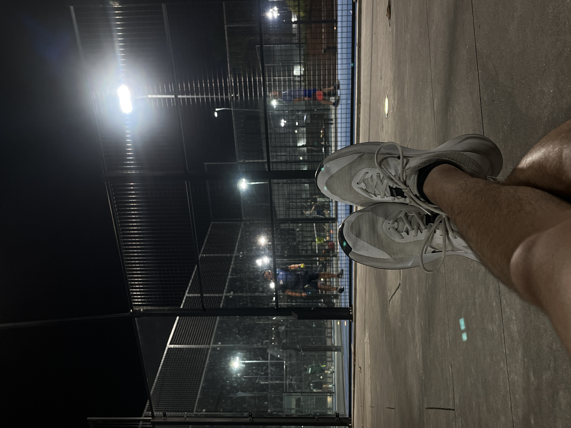 The image presents a first-person perspective from the edge of a sports court at night. In the foreground, a person's bare, hairy legs are visible, extending forward with feet encased in white athletic shoes featuring prominent laces. These feet rest on a textured grey surface, likely concrete or similar court-side flooring, which reflects the bright artificial lights.
The main subject is a padel or tennis court, enclosed by tall black wire mesh fences and clear glass panels. The scene is brightly illuminated by numerous artificial lights, casting reflections on the court surface and glass, starkly contrasting with the dark night sky visible beyond the structures. Several blurry figures are discernible within the court, actively engaged in a game, indicating an ongoing match or practice session. One player, wearing a blue shirt and red shorts, is somewhat clearer in the mid-distance, while another player in a green shirt is visible further back. The image conveys the atmosphere of an evening sports event or recreational play. No visible text is identifiable.