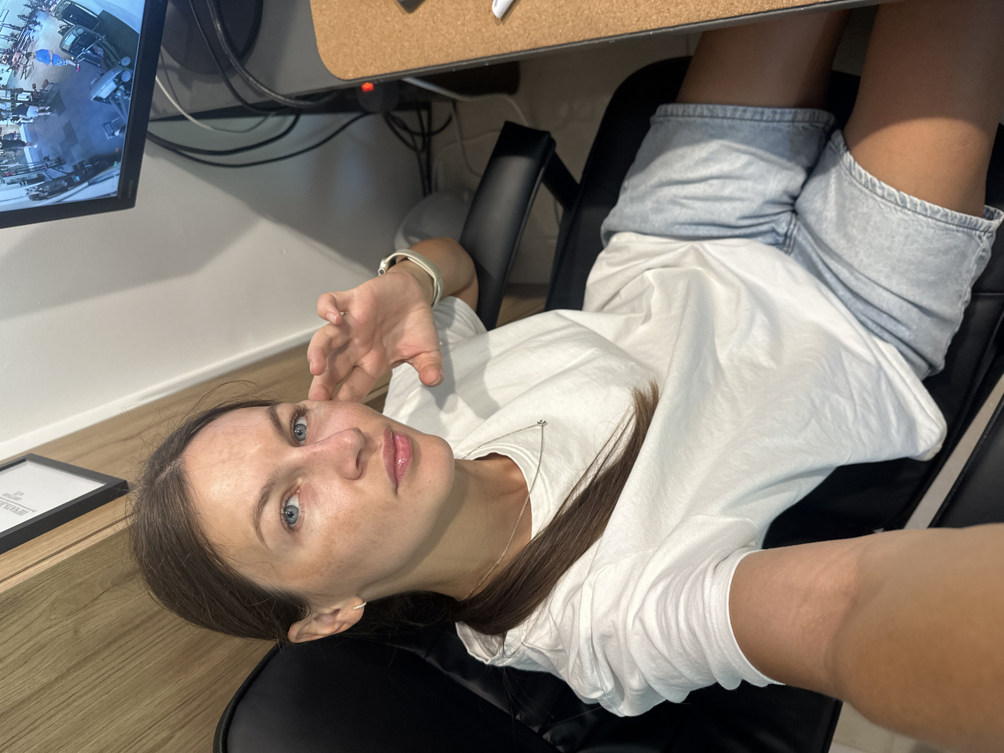 A woman is sitting in an office chair and looking at the camera. She has brown hair, blue eyes, and fair skin with freckles. She is wearing a white t-shirt and denim shorts. She is sitting at a desk with a computer monitor in front of her. The monitor shows a view of a gym. The room appears to be an office or home office, with a wooden wall behind the woman and a cork-covered desk to her right. The lighting is bright, suggesting it is daytime. The woman's pose and gaze suggest she is aware of being photographed. The image is taken from a slightly low angle, looking up at the woman.