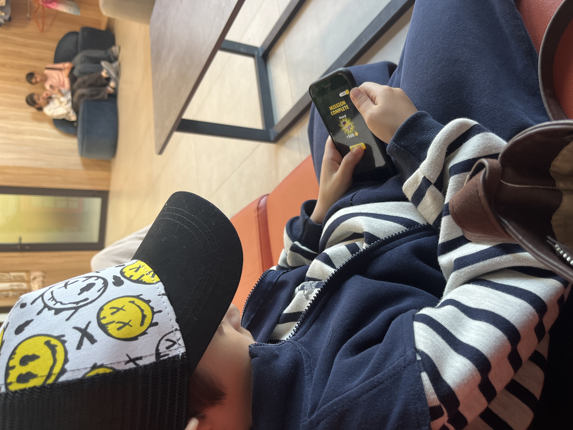 A person wearing a black and yellow smiley face cap is lying on an orange seat, looking at a phone that displays "Mission Complete". The person is wearing a navy blue jacket with white stripes. The location is Kaspiysk, Russia. Other people are sitting in blue chairs in the background. A table is visible between the person and the chairs. A purse is next to the person.