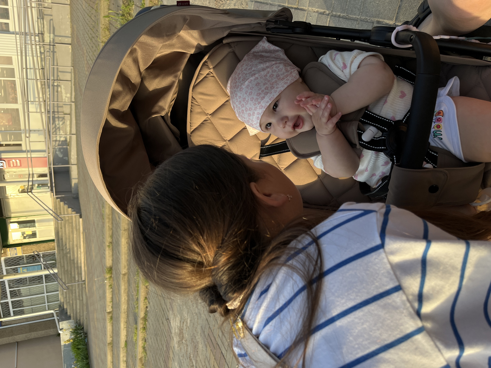 A young child wearing a pink floral head covering sits in a tan stroller and faces a person wearing a white shirt with blue stripes, who is looking down at the child. The stroller is positioned on a sidewalk. In the background, there is a building and a set of stairs. This scene appears to be occurring in the city of Novopokrovskaya, Russia.