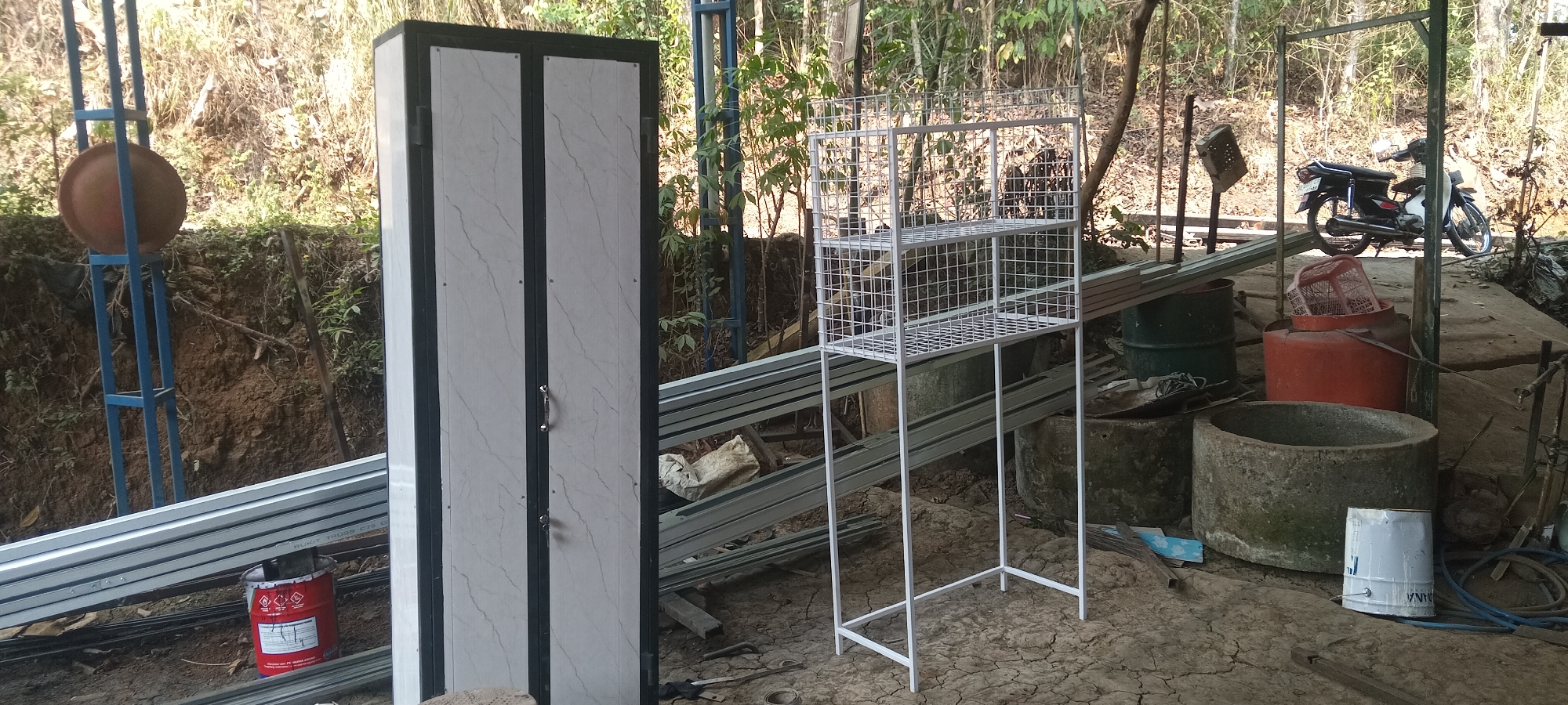 The image depicts an outdoor storage area, likely in a rural setting, with a focus on metal shelving units and construction materials. The scene is dominated by a tall, narrow cabinet with two doors, its exterior patterned with a marble-like design. To its right stands a white metal rack with three open shelves, constructed from wire mesh. In the background, a black and white motorcycle is parked. The ground is covered in cracked, dry earth. Various large cylindrical objects, some appearing to be drums or concrete structures, are scattered throughout the area, along with piles of metal beams and a red paint can. Lush green vegetation and trees form a backdrop, suggesting an outdoor environment. The overall impression is one of a workspace or storage facility for construction or home improvement materials.