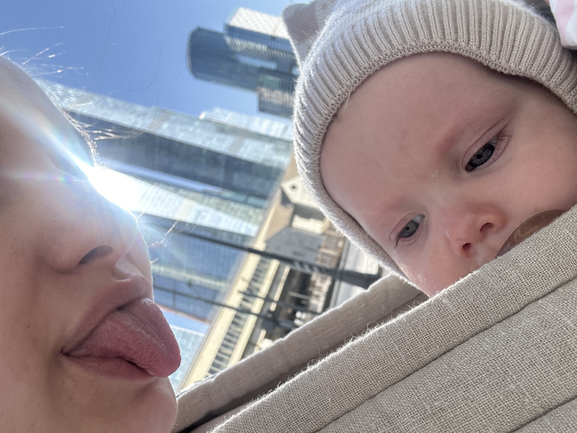 The image depicts a close-up, outdoor scene in an urban environment near Khimki, Russia. The upper-left portion of the frame shows the lower face of an adult, specifically the nose and mouth, with a pink tongue fully extended. Below and to the right, an infant is visible, partially swaddled in a light beige, textured fabric, consistent with a baby carrier or wrap. The infant wears a light-colored, ribbed knitted cap with two ear-like protrusions and has fair skin and blue eyes. A brown pacifier is partially visible in the infant's mouth, and its gaze is directed slightly off-camera to the left. The background features a clear blue sky and several modern glass-facade skyscrapers, characteristic of the Moscow City area, directly adjacent to Khimki. An elevated concrete structure, possibly a bridge or overpass, is discernible below the skyscrapers. Intense sunlight causes a prominent lens flare in the upper-middle section of the image.