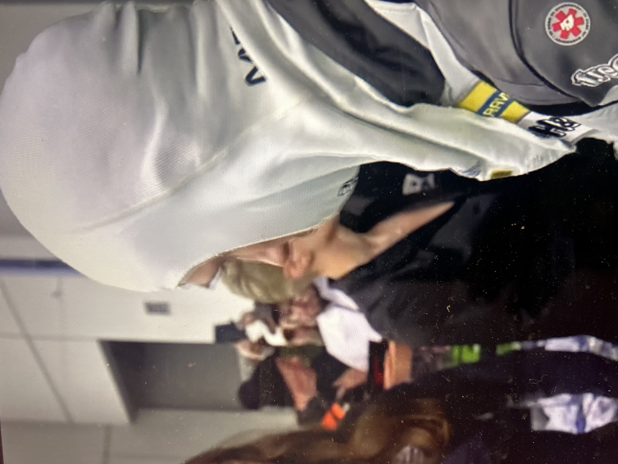 The image shows a person wearing a white hooded sweatshirt and a black jacket with a patch featuring a red cross design and lettering. A yellow band with black text (likely "РАВ") is visible on the jacket's sleeve. Other individuals are blurred in the background. The image is of poor quality. There is no discernible information in the image that would reveal the city of Sofia, Bulgaria as the location.
