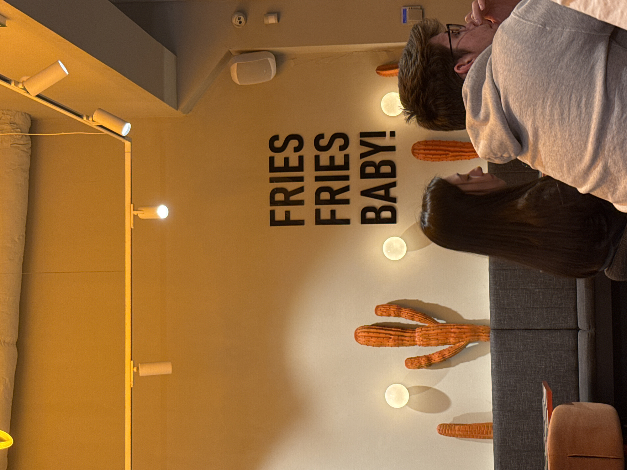 The image captures an interior scene. A wall is adorned with the text "FRIES FRIES BABY!" and decorative cactus-shaped objects illuminated by round lights. To the right, a man with glasses wearing a gray hoodie stands next to a woman with long dark hair, partly obscuring the wall. A gray sofa is visible near the woman, and a glimpse of a reddish-brown chair is at the bottom right corner. The scene is brightly lit, with additional track lighting on the left side of the frame. The location is Sofia, Bulgaria.