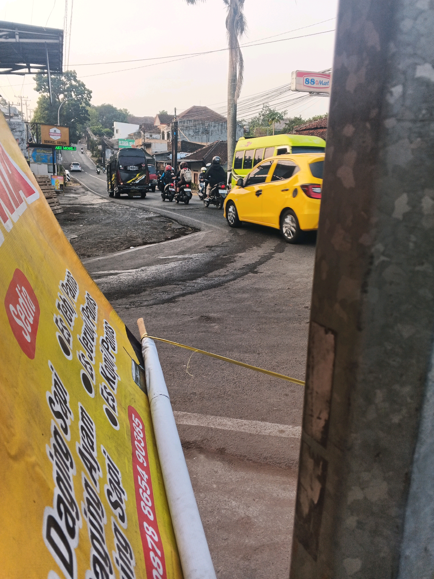 The image depicts a street scene in Semarang, Indonesia. In the foreground, a large yellow banner with Indonesian text and graphics is partially visible, angled towards the viewer. The text on the banner appears to be a menu or advertisement, with words like "Sedia," "Daging Sapi," and "Ayam" (beef, chicken).
The street is paved and appears to be a road with a slight incline. Several vehicles are present, including a black bus with colorful graphics, a bright yellow car, and multiple motorbikes with riders. Some of the motorbikes have passengers. The vehicles are moving along the road, suggesting it is a relatively busy street.
In the background, there are buildings, some of which appear to be houses with traditional roofs. A tall palm tree is visible on the right side of the frame. There are also power lines and poles, indicating urban infrastructure. A sign for "88 Mart" is visible overhead, suggesting a commercial area. The overall lighting suggests it might be late afternoon or early morning, with a soft, diffused light. A metal pole is prominently positioned in the far right of the image, partially obscuring the view.