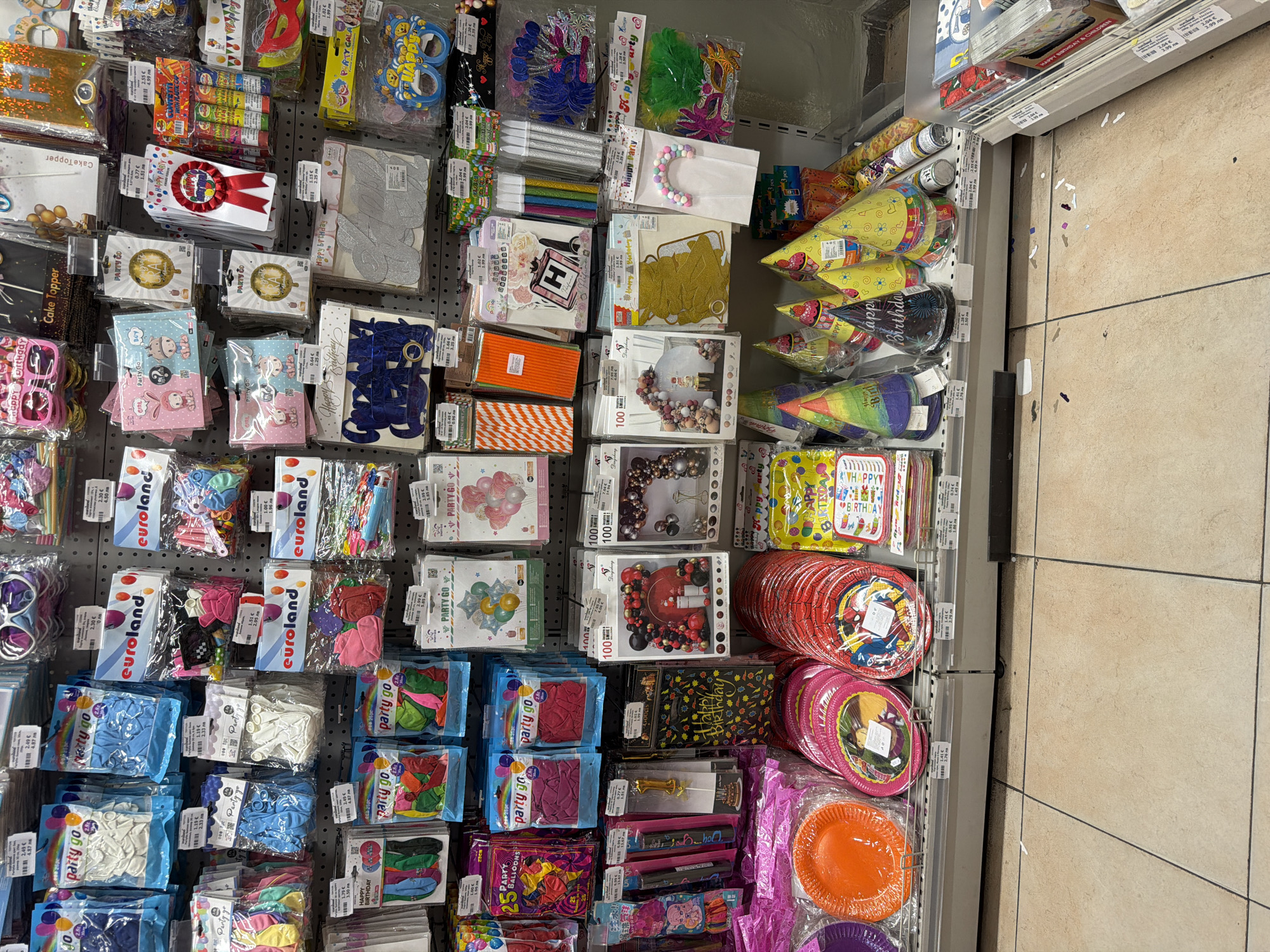 The image shows a retail display of party supplies, specifically for birthdays. The shelves are stocked with items such as balloons, paper plates, party hats, banners, and other decorations. The items are mostly packaged and neatly arranged. In the foreground, the tiled floor of the store is visible, with some scattered confetti and small pieces of paper. The overall impression is of a well-organized party supply store. The location of Pazardzhik, Bulgaria is not visible in the image, as the image is a close-up of a retail display.