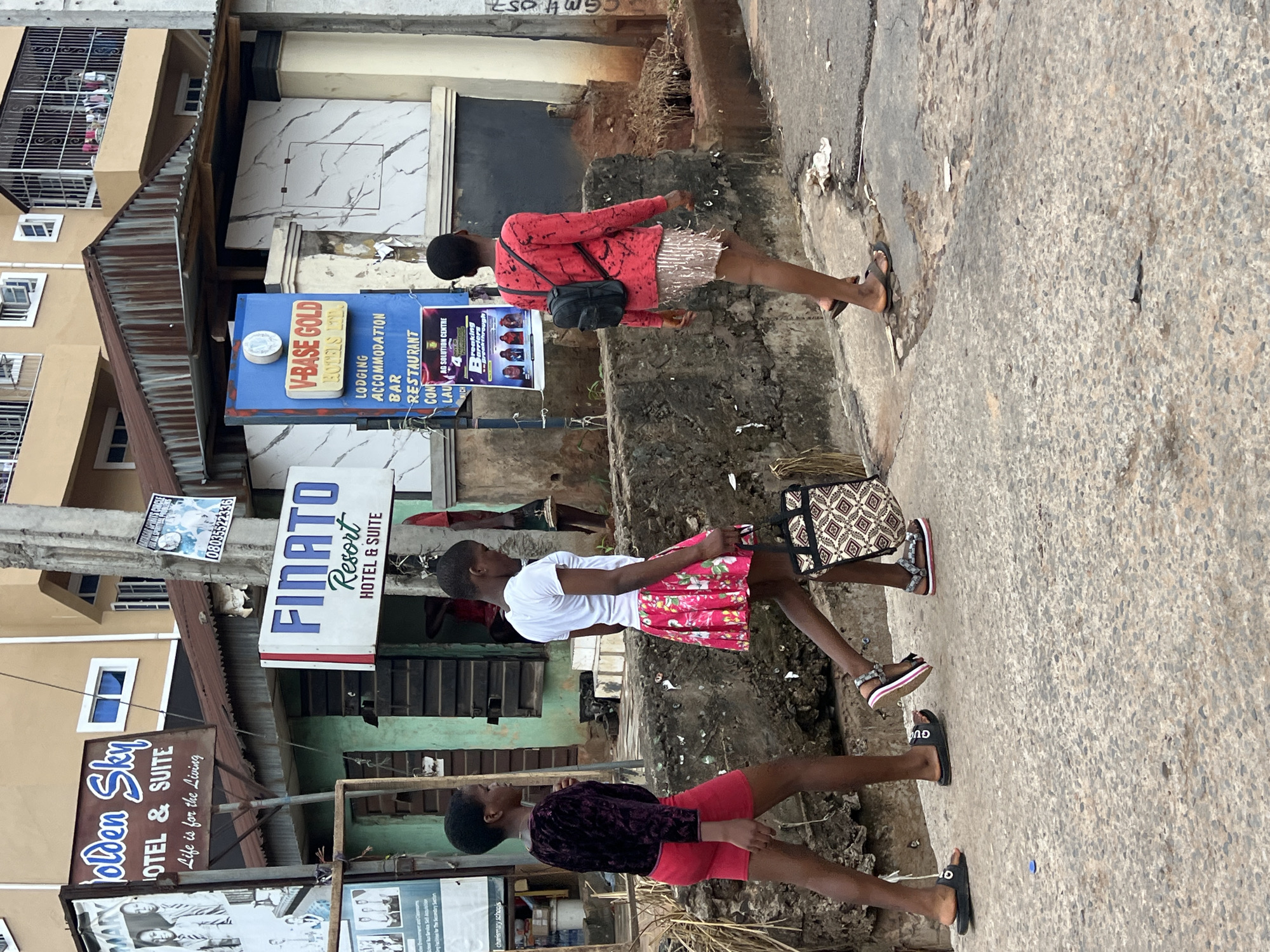 In Nkpor, Nigeria, three young girls walk along a concrete path beside a street. The girl on the left wears a dark velvet long-sleeved top and a red skirt. She is wearing black flip-flops. The girl in the middle has on a white t-shirt and a floral pink skirt. She is wearing sandals and carrying a tote bag with a geometric pattern. The girl on the right is wearing a red long-sleeved shirt and a sparkling silver skirt. She has a black backpack on and is wearing brown flip-flops. They are walking past buildings with signs, including "Golden Sky HOTEL & SUITE" and "FINATO Resort HOTEL & SUITE." A blue sign for "V-BASE GOLD HOTELS LTD." is also visible, listing lodging, accommodation, bar, and restaurant services. The environment appears somewhat weathered, with a corrugated iron roof visible on one of the buildings and rough concrete textures.