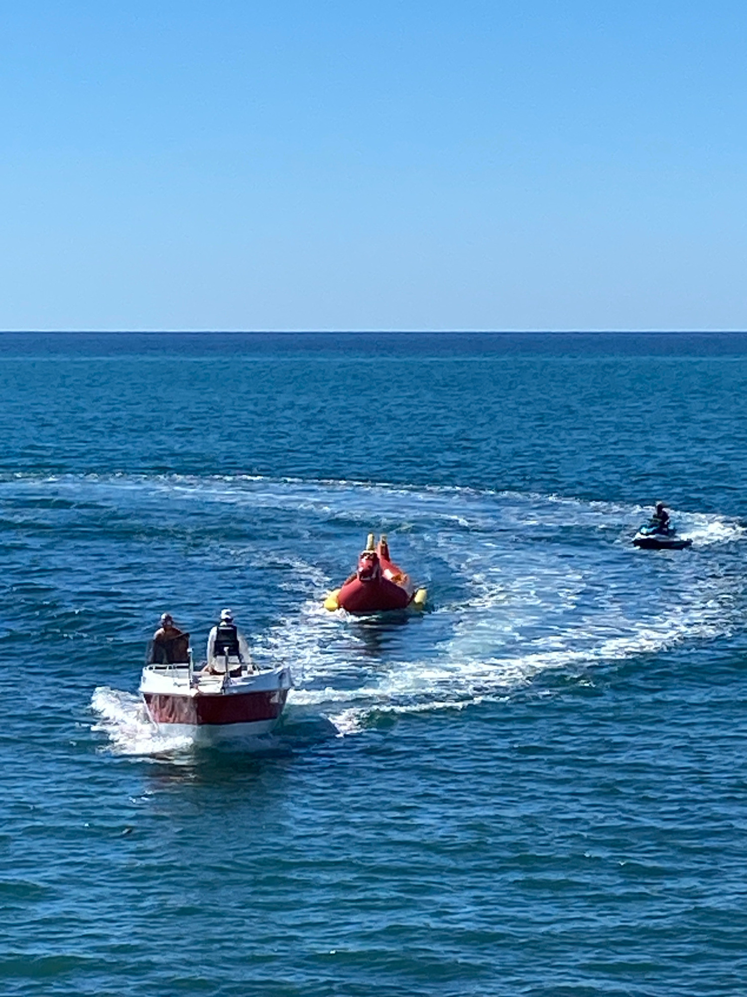 In the city of Sochi, Russia, a bright blue sky meets the deep blue water of the sea. Three recreational watercraft are in motion, leaving white wakes behind them. In the foreground, a small white and red motorboat carries two individuals. Further back, a person is being pulled on a red inflatable tube, lying on their back with their legs in the air. To the right, a jet ski is being ridden by a single person, also creating a visible wake. The scene appears to be a sunny day with calm seas.