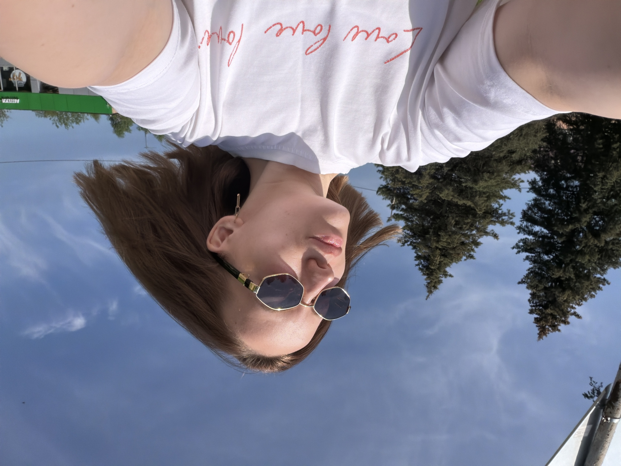 A woman wearing sunglasses and a white t-shirt with red lettering is the subject of this selfie. The backdrop reveals a clear, blue sky with sparse clouds and the tops of two evergreen trees. In the upper left corner of the frame, a green sign indicates a business or location, "ARTEK." The location appears to be an outdoor space in Reshetnikovo, Russia.