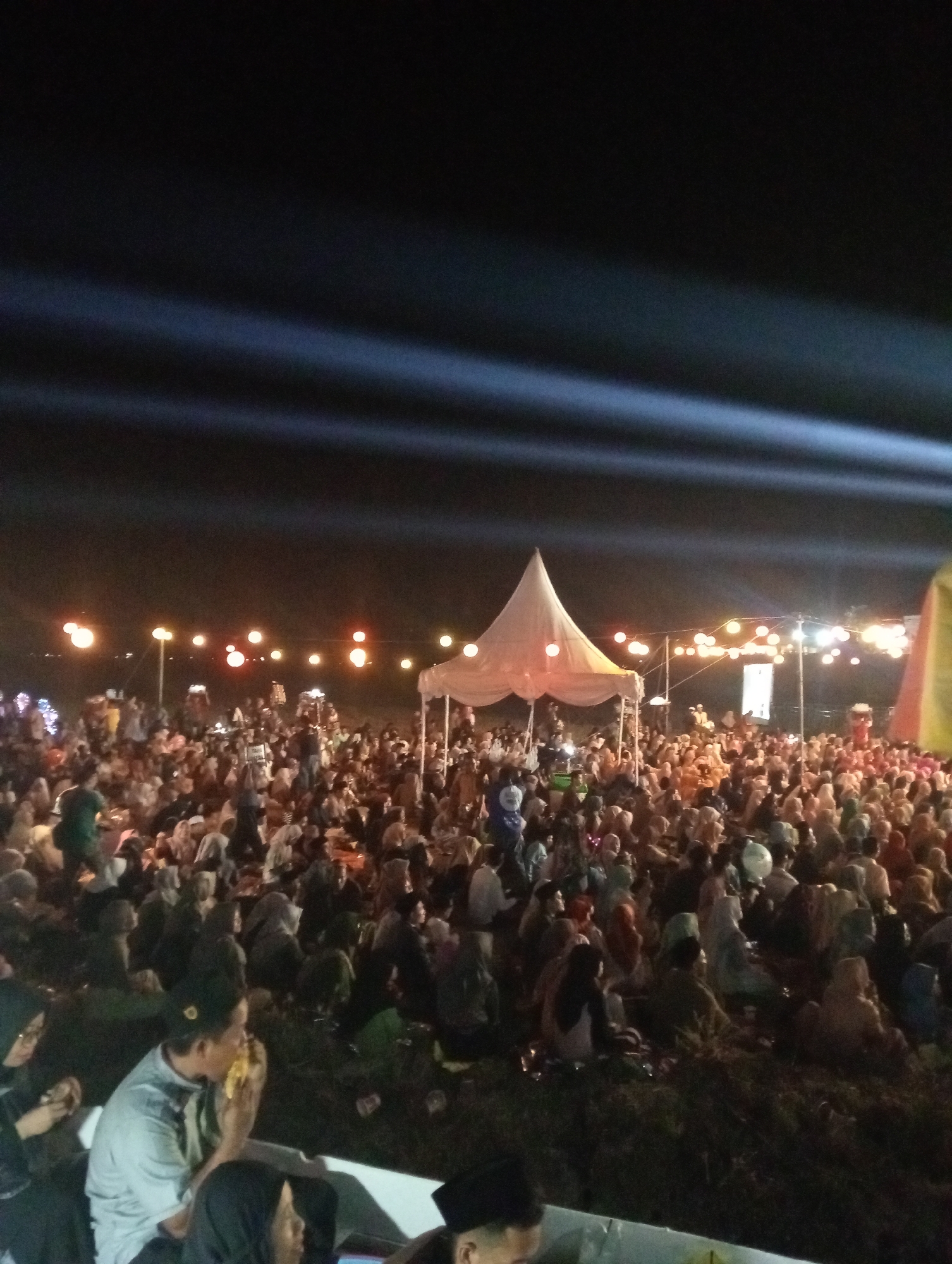 The image depicts an outdoor night event with a large crowd gathered, possibly in the city of Wonorejo, Indonesia. A white, peaked tent is centrally located, and string lights with warm yellow and orange bulbs illuminate the scene. In the background, bright white beams of light are projected across the dark sky, suggesting a performance or stage lighting. Many people are seated on the ground, facing towards the tent and the illuminated area, indicating they are attending an event such as a concert, religious gathering, or festival. The attendees appear to be dressed in casual attire, with some individuals wearing headscarves. The overall atmosphere seems festive and communal.