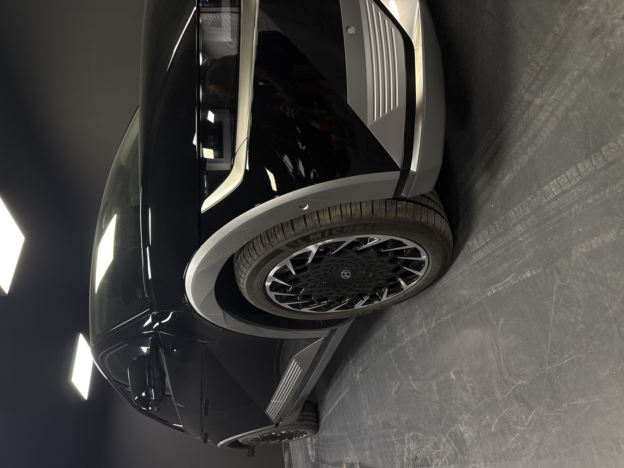 A black Hyundai Ioniq 5 is parked indoors, likely in a garage or showroom, given the dark walls and ceiling with overhead lighting. The car is positioned at an angle, showcasing its front-right side. The vehicle features distinctive pixelated headlights and taillights, a design element that defines its modern aesthetic. The wheel arches are accented with a contrasting gray trim, and the car is fitted with black alloy wheels featuring a complex, multi-spoke design. The floor beneath the car is a dark, industrial-looking surface, marked with tire tracks and scuffs, indicating it's a functional space. The overall lighting is bright and artificial, casting shadows that highlight the car's sharp lines and reflective surfaces. There are no people clearly visible in the image, but reflections in the car's paint suggest there might be individuals present, possibly taking the photograph or observing the vehicle. There is no indication that this scene is located in Ruse, Bulgaria, or any other specific city.