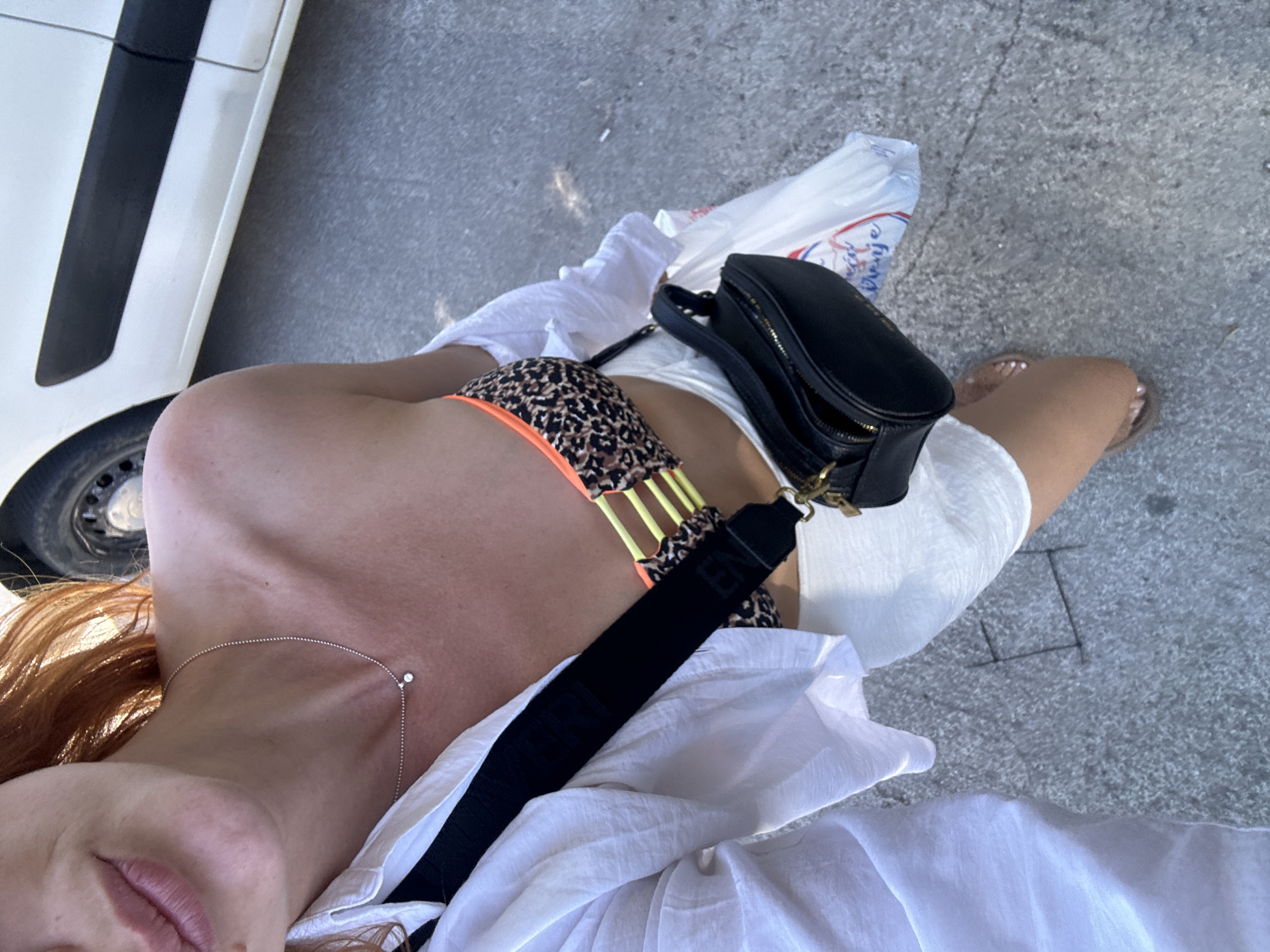 A woman with red hair is taking a selfie outdoors. She is wearing a white open button-up shirt over a leopard-print bikini top with yellow accents, and white shorts. She has a small silver necklace with a diamond pendant. A black crossbody bag with gold hardware is slung across her body. She is holding a white plastic shopping bag in her right hand. Her feet are visible at the bottom of the frame, wearing flat sandals. The background shows a paved surface and a white vehicle with a black stripe. The scene is likely in Budva, Montenegro, given the context provided, perhaps near the coast or a street with shops.