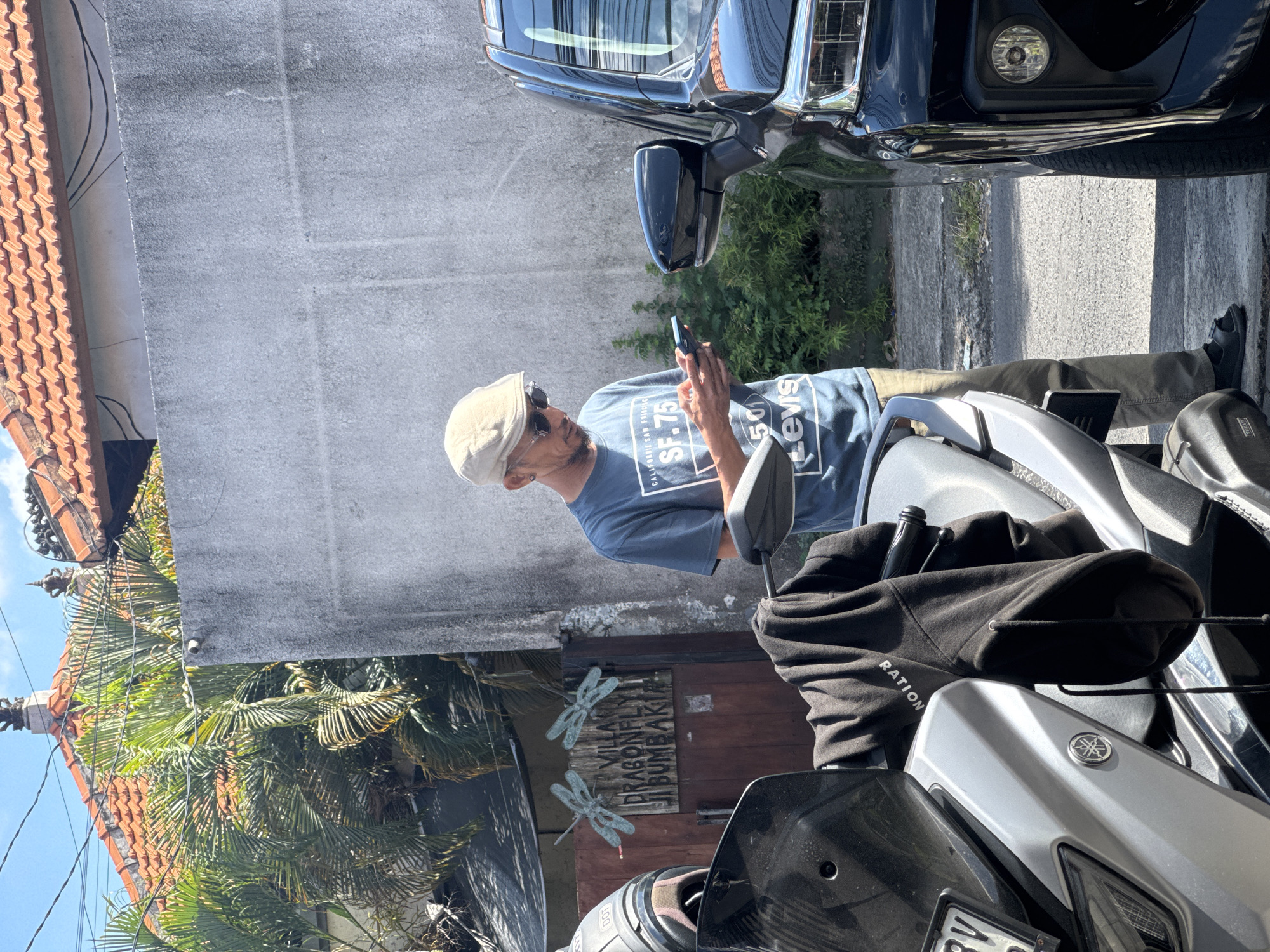 The image captures an outdoor scene in bright daylight, likely in North Kuta, Indonesia. A man, appearing to be in his 30s or 40s, stands next to a silver and black scooter, intently looking down at a dark smartphone held in both hands. He wears a light-colored flat cap with sunglasses resting on top, a blue t-shirt with "CALIFORNIA SAN FRANCISCO SF.75 LEVIS" and "501" printed on it, khaki pants, and dark shoes. He has a dark beard.
In the immediate foreground, the front part of a scooter, identifiable by a Yamaha emblem, is visible. A black hoodie, partially revealing "RATION" text, is draped over its handlebars or seat, with a black bottle visible inside. A license plate partially reads "DK 5V8". Behind the man, a dark blue or black car is parked, showing its front grille, headlight, and side mirror.
The background features a textured grey concrete wall. To the left, lush green tropical foliage, including palm fronds, is visible, partially obscuring a traditional red-orange tiled roof with decorative elements. A wooden door beneath the foliage is adorned with two metallic dragonfly decorations and a carved sign that reads "VILLA DRAGONFLY UBUD BUMBAK". The bright, direct sunlight casts strong shadows, indicating a clear, sunny day.