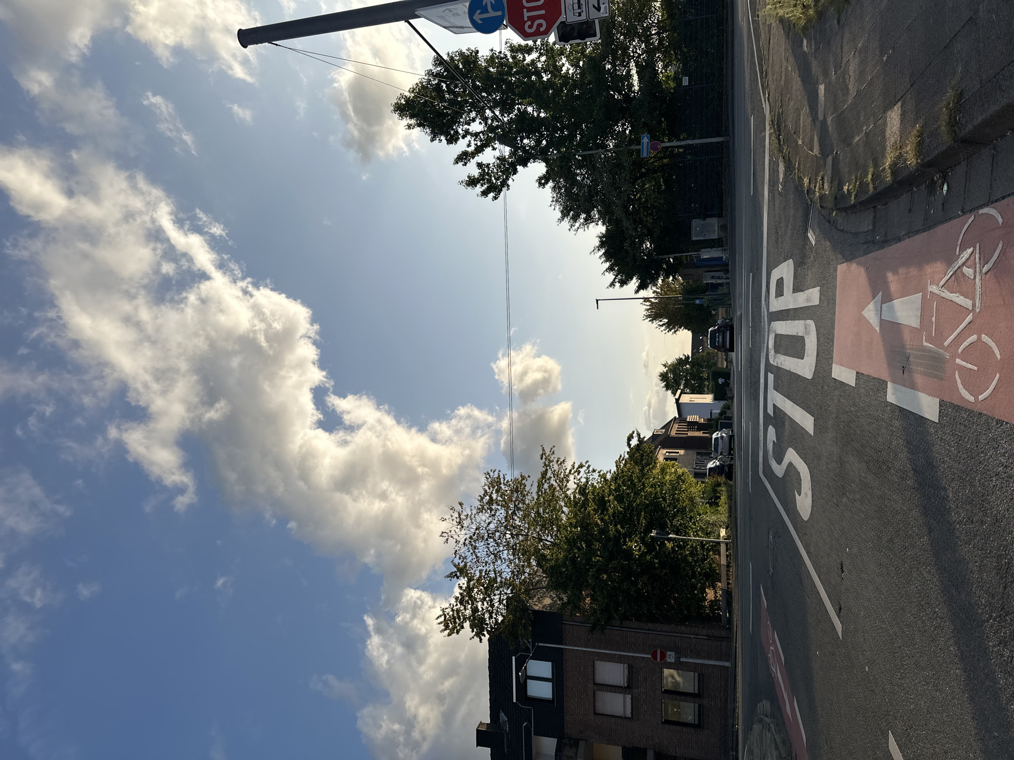 The image depicts a street intersection in Krefeld, Germany, under a sky with scattered clouds and the late afternoon sun casting long shadows. The foreground features an asphalt road with painted markings for a bicycle lane, indicated by a red background and a white bicycle symbol with an arrow pointing forward. A white "STOP" sign is painted across the lane.
To the left, a multi-story brick building with dark-tiled roofing and white-framed windows is visible. Trees with green foliage are situated between the building and the road. On the right side of the intersection, more trees and buildings are present, as well as street signs. One prominent sign is a red octagonal STOP sign, accompanied by a blue circular sign with a white arrow indicating a right turn and a straight path. Another sign shows a tram symbol and an arrow pointing to the right.
Several cars are parked or driving in the background, and a few individuals are faintly visible walking on the sidewalk or near the buildings. The overall impression is a typical urban street scene with clear traffic control elements.