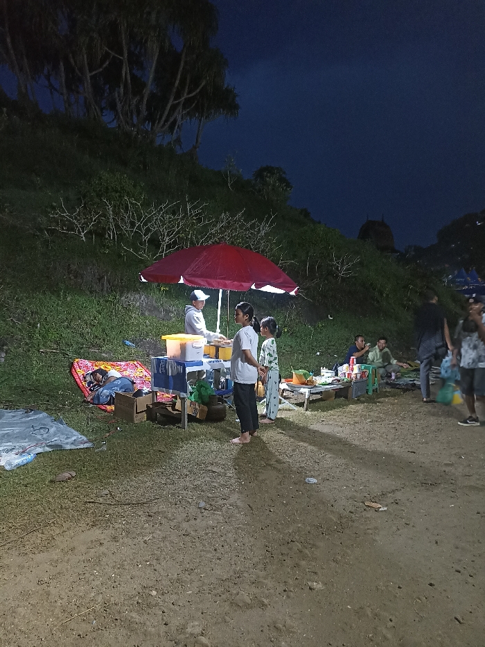 This night-time image captures an outdoor scene, likely a temporary market or food stall, in Kecamatan Pujut, Indonesia. The central focus is a vendor, a man in a white cap and hoodie, standing behind a makeshift stall with yellow and blue containers under a bright red umbrella. Two young girls, one in a white t-shirt and black pants, and another in patterned pajamas, stand before him, seemingly engaged in a transaction or viewing items.
To the left of the vendor's stall, a person is asleep on a red mat laid on the grass, covered by a blanket. Nearby, a blue and white patterned cloth or blanket lies on the ground. To the right of the stall, several other individuals are visible. Some are seated on small stools around a low table laden with various items, possibly food and beverages, while others stand further back. One man on the far right wears a light-colored shirt and patterned shorts.
The ground is unpaved and earthy, transitioning to a grassy slope densely covered with bushes and trees in the background. The dark sky confirms it is nighttime, with artificial lighting illuminating the immediate area around the stall and people, casting distinct, elongated shadows on the ground. The scene depicts a casual gathering with people socialising and accessing refreshments in an informal outdoor setting. No legible text is visible in the image.