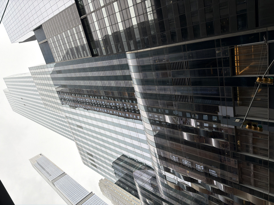 The image displays a low-angle, upward view of multiple skyscrapers in Midtown Manhattan, New York City, United States of America, under an overcast sky.
The central focus is on two prominent high-rise buildings. To the left, a very tall, slender skyscraper, identifiable as 432 Park Avenue, features a facade composed of light-colored, square-patterned glass panels. Adjacent to it on the right, another significant skyscraper, known as 53W53 (MoMA Tower), exhibits a darker, more intricate facade with a distinctive undulating or wavy pattern of reflective glass and steel, particularly visible in its lower and mid-sections. This building reflects parts of the surrounding architecture and the sky. To the far left, another tall, gridded glass tower is partially visible. To the right of 53W53, a darker building with a less reflective, grid-patterned facade and opaque sections rises.
The lower portion of the image includes structural elements of the buildings and, at the bottom right, two sets of three-light traffic signals are suspended on an arm, displaying green lights. No individuals are visible. The scene is devoid of specific actions or interactions, solely presenting architectural forms in a dense urban environment.