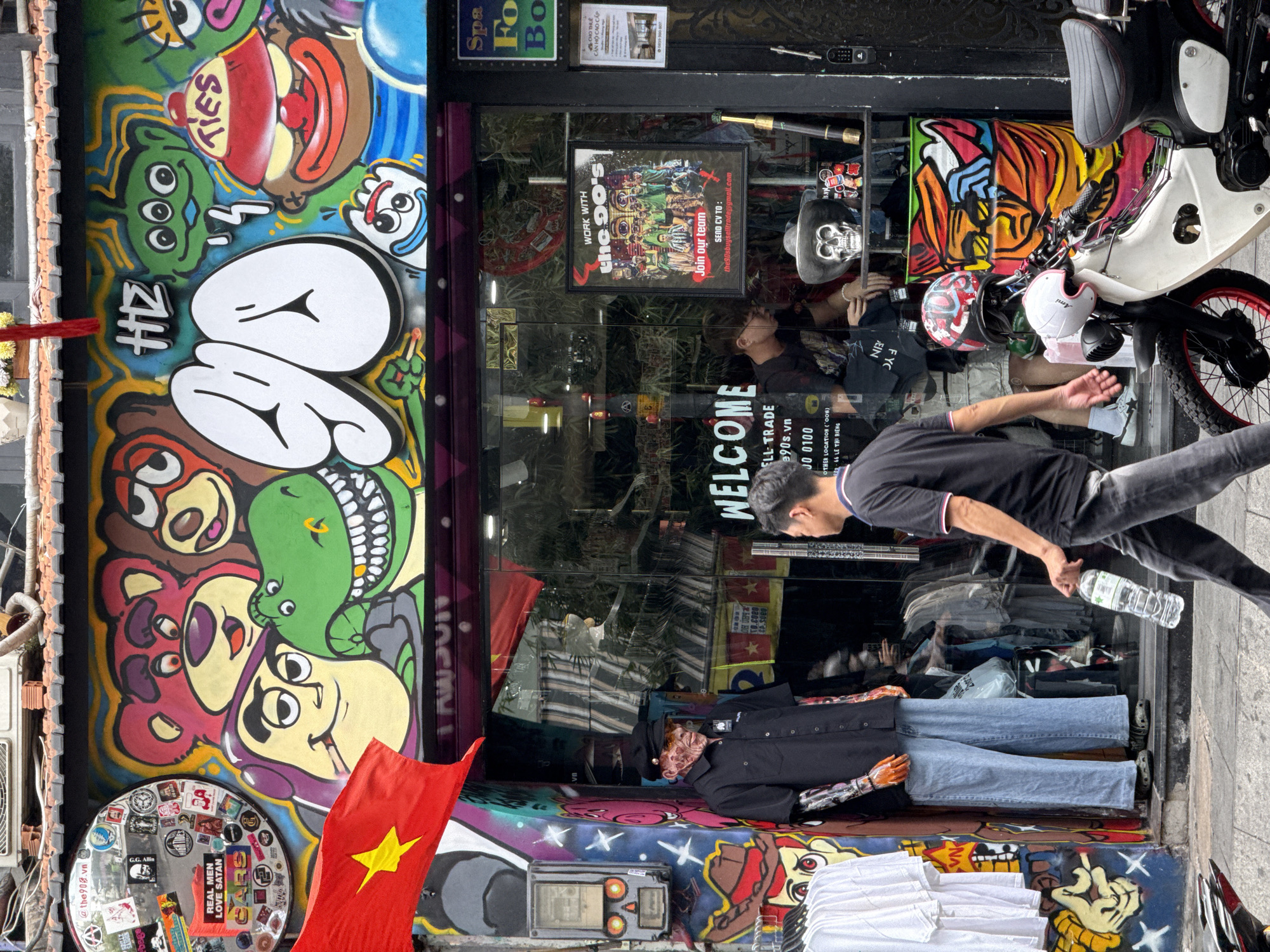 The scene is an outdoor street-level view of a storefront, possibly a clothing store in Thủ Đức, Vietnam. The building exterior features vibrant graffiti art depicting cartoon characters and designs. A Vietnamese flag is visible on the left side. A man walks by the storefront holding a water bottle. Inside the store window, there's a mannequin dressed in a black shirt and jeans, arranged horizontally on a shelf with folded white shirts nearby. There's a "Welcome" sign in the window along with contact information and the label "ELL TRADE." Several other elements are visible through the glass, including a motorcycle with helmets, a skull with a hat, and posters, including one advertising work for "the 90's." There's a sticker-covered round object (possibly a mirror or sign) attached to the wall on the left. The scene appears to be captured during daylight.