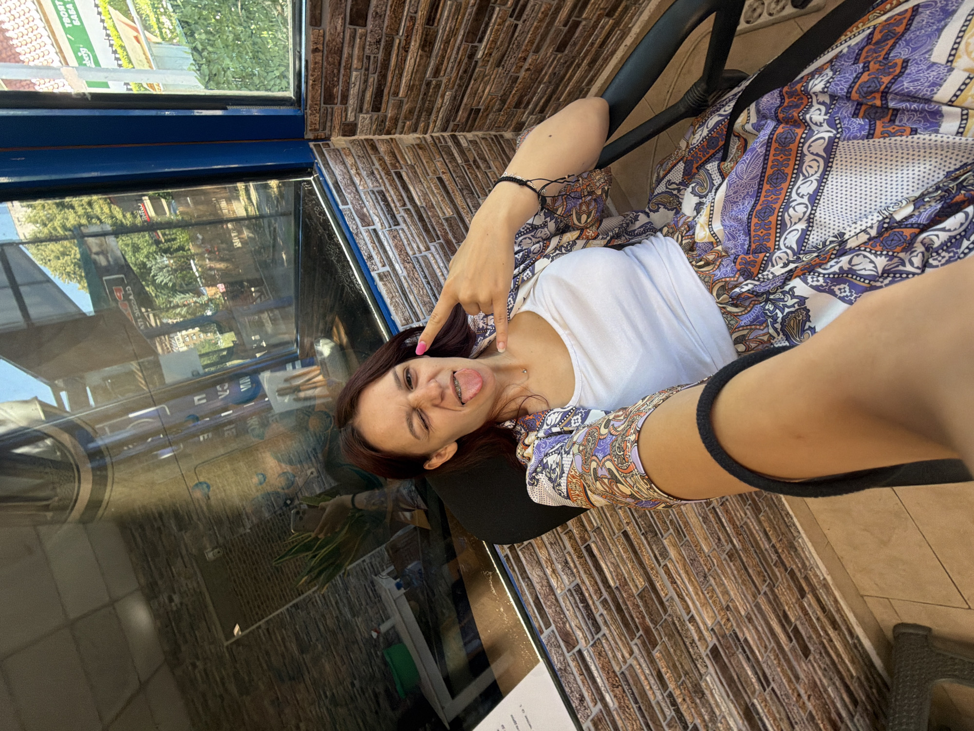 The image shows a young woman with reddish-brown hair sitting in an office chair, taking a selfie with her phone. She is wearing a white tank top and a patterned skirt and blouse. She is winking and sticking out her tongue, with her right hand making a rock-and-roll gesture. The background appears to be a cafe or restaurant with a large window reflecting the exterior. The wall behind the woman is made of stacked brown bricks. Through the window, trees and buildings are visible, suggesting an urban outdoor setting. The cafe appears to be located in Sofia, Bulgaria, as indicated by the text visible in the reflection on the window, which includes Cyrillic script and a logo for "Berg."