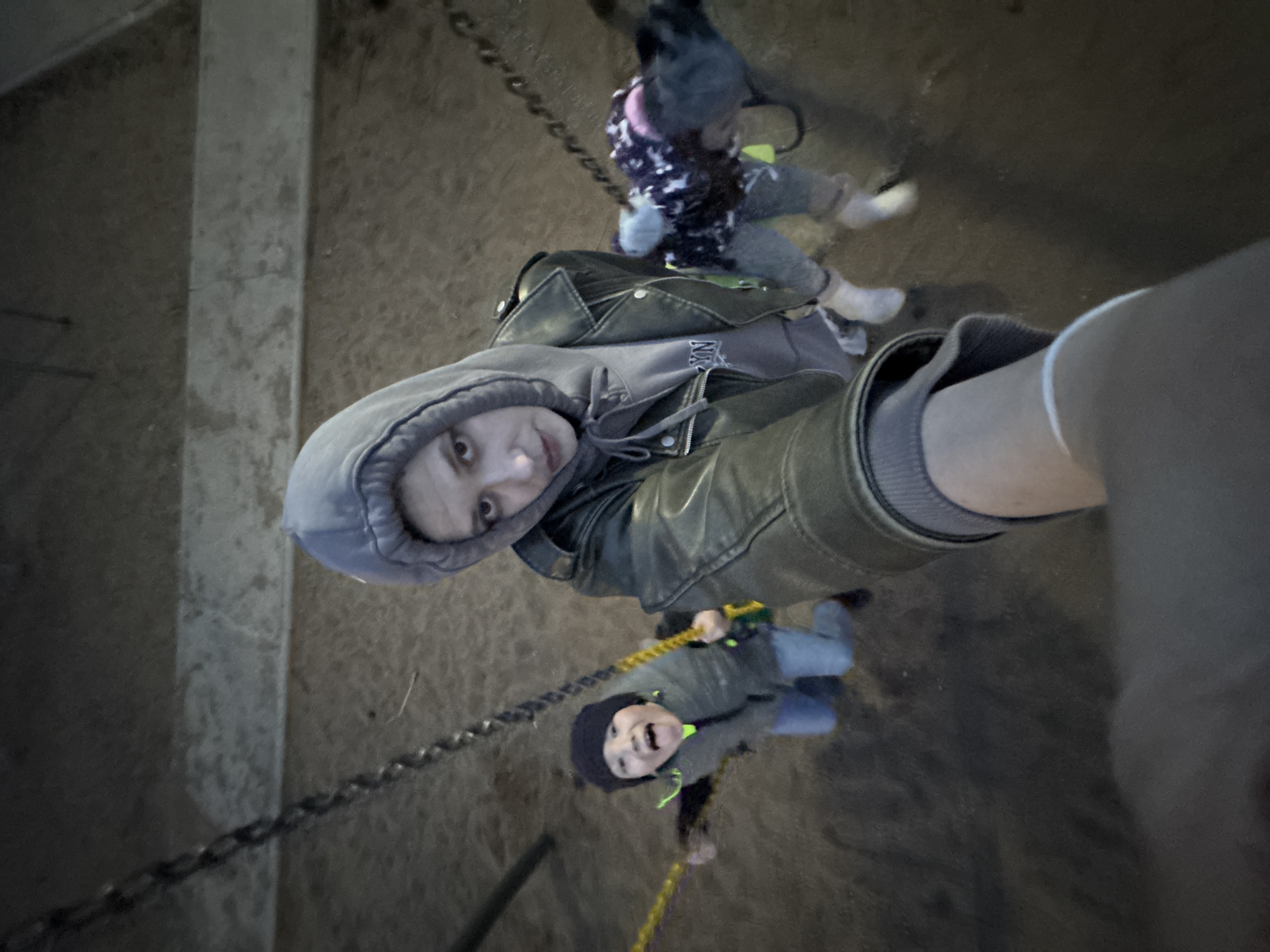 The image depicts a selfie taken from a high angle, showing a person wearing a hooded jacket and two young children. The children are on swings, held up by chains. The background appears to be an outdoor playground with a surface that looks like sand or dirt. The location is in Chaikovsky, Russia.