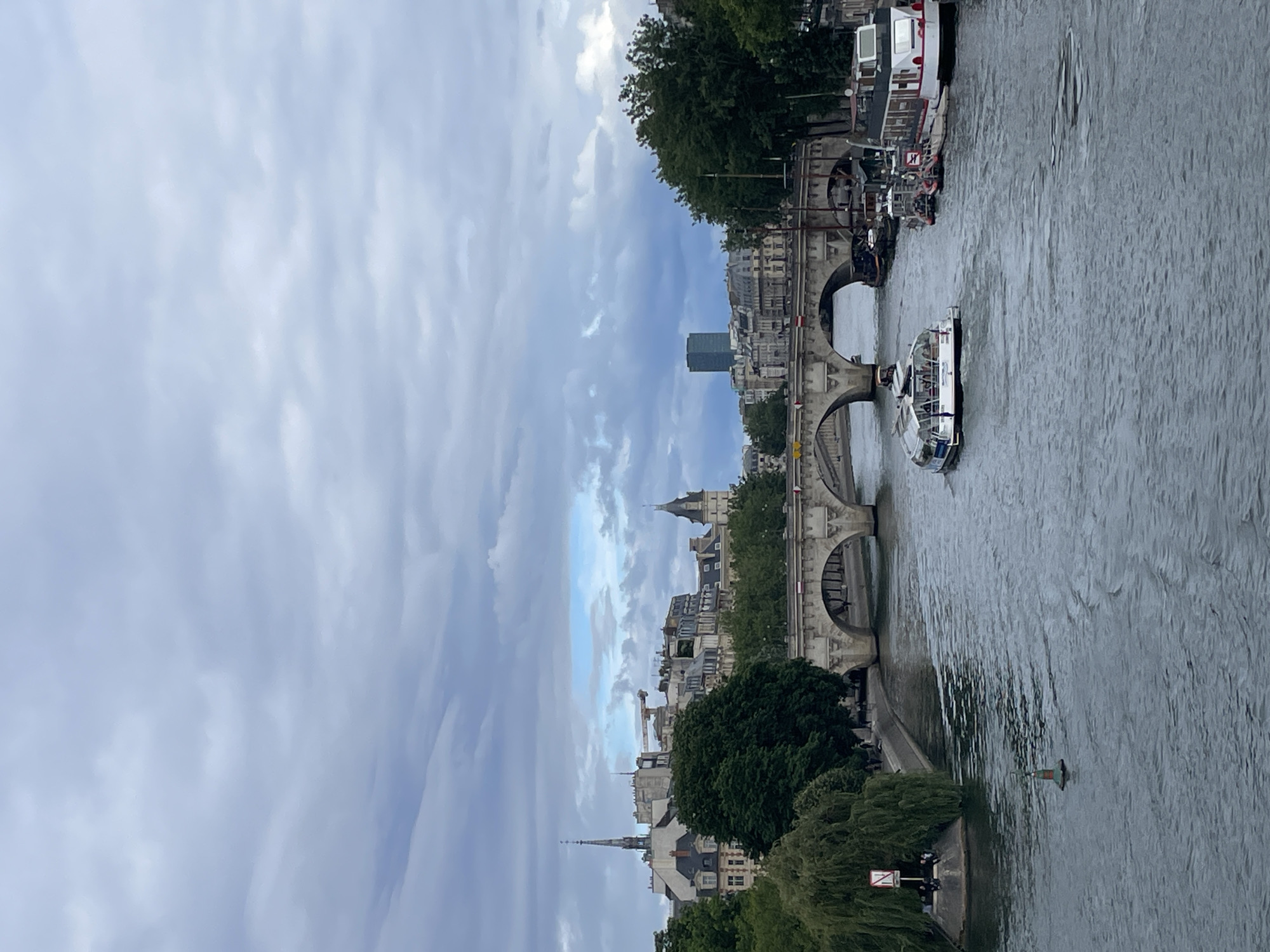 The image captures a view of the Seine River in Paris, France, under a cloudy sky. A modern tourist boat with large windows is moving downstream, while several other boats, including a houseboat, are docked along the right bank. A stone bridge with multiple arches spans the river in the middle ground, with buildings and trees lining the riverbanks on either side. The architecture suggests a historical European city. The weather appears overcast, casting a diffused light over the scene.
