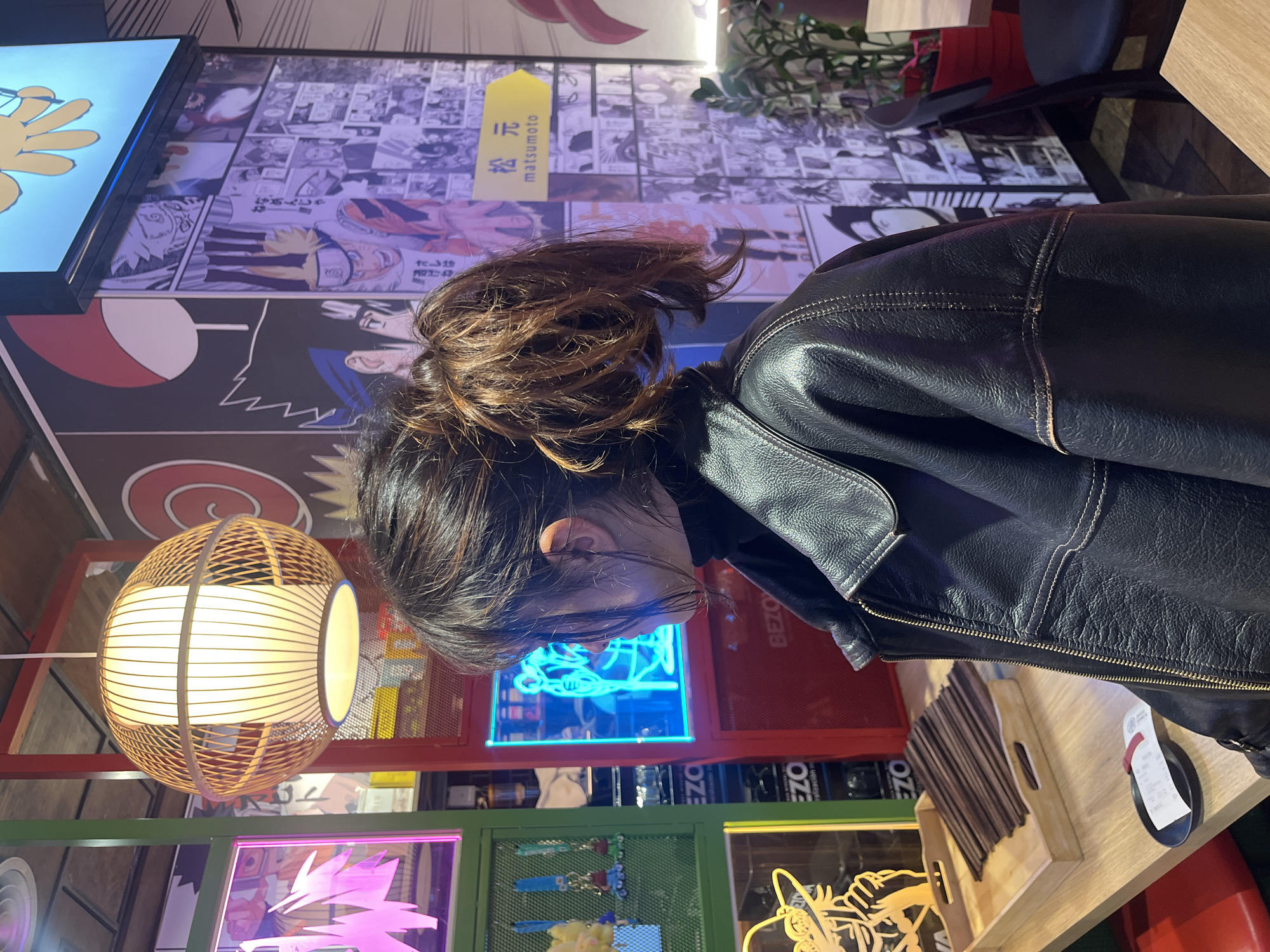 A medium shot reveals a person with dark hair and a black leather jacket sitting at a table. The jacket's texture is visible. The person is looking downwards, with their face partially obscured by their hair. The background features a wall decorated with manga-style drawings and a television screen. A round, caged lamp hangs nearby. Below, colorful neon signs and displays line a shelf, suggesting an urban environment, possibly a restaurant or shop in Barcelona, Spain. The table has chopsticks and a payment tray.