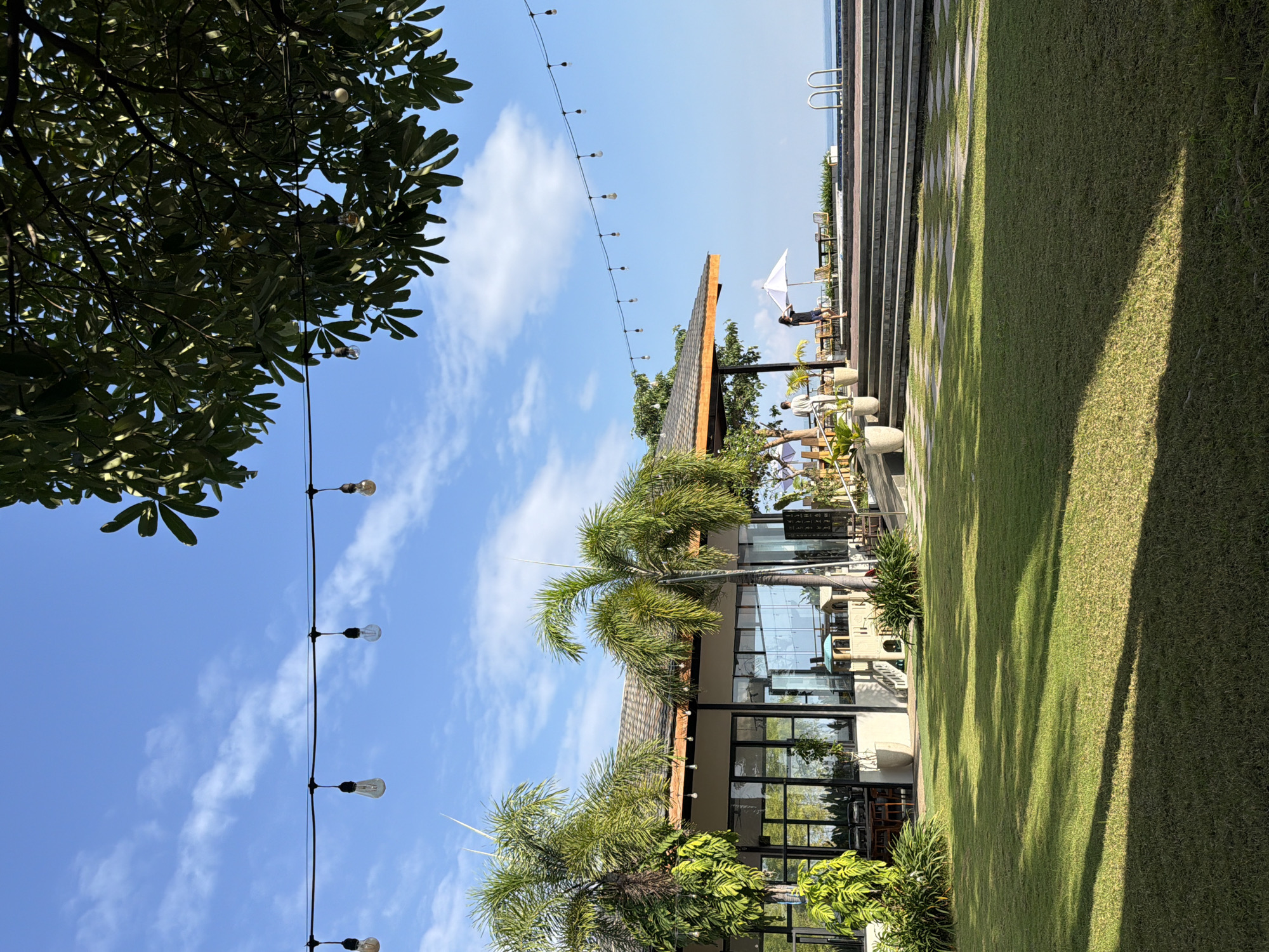 A resort restaurant with a lawn and ocean view is shown in Pasir Putih, Indonesia. The restaurant has large windows and is decorated with string lights. There are palm trees and a playground on the property. In the foreground, a lush green lawn is illuminated by sunlight, casting shadows across the grass. A woman in a dark dress is standing near a white umbrella, while another person in light clothing stands nearby. The ocean is visible in the background, beyond a tiled patio area with seating.