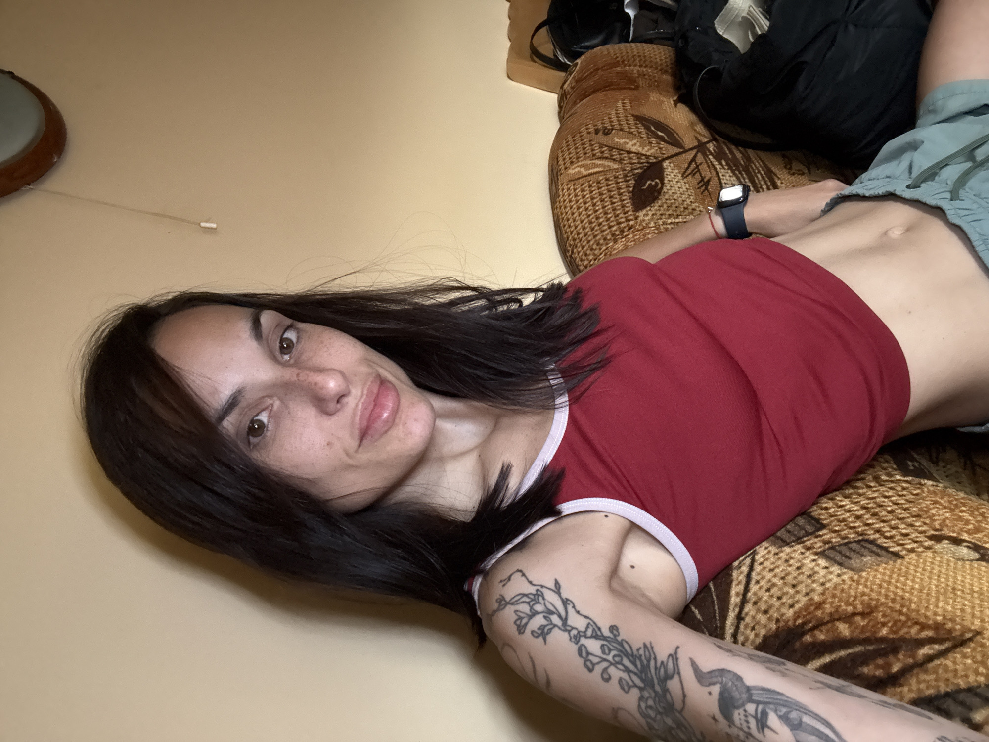 The image shows a young woman with dark hair and freckles, wearing a red tank top and green shorts. She has tattoos on her left arm and is wearing a watch on her left wrist. She is sitting on a couch with a patterned fabric. The background is a plain, light-colored wall. There is a light fixture visible on the wall. The image appears to be taken indoors. There is no indication of the city of Pleven, Bulgaria.
