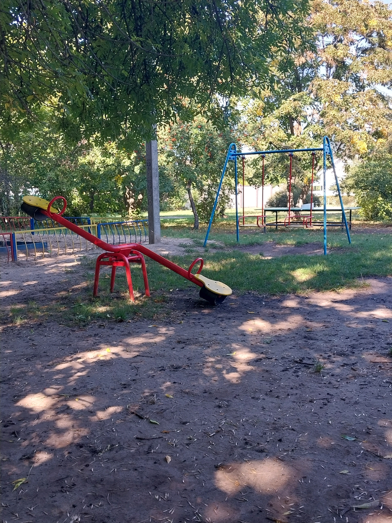 The image depicts an outdoor playground area in the village of Doslidne, Ukraine, on a sunny day. In the foreground, a red seesaw with yellow seats is positioned on a dirt ground covered with scattered leaves and grass. To the left, a blue and yellow metal fence surrounds a sandbox. In the background, a blue metal swing set with red swing seats is visible. Behind the swing set, there is a picnic table with benches. Mature trees with green and yellow foliage provide shade over the playground. A concrete pole stands near the center of the image. The ground is uneven and appears to be a mix of dirt, grass, and fallen leaves. Sunlight casts dappled shadows from the trees onto the ground.