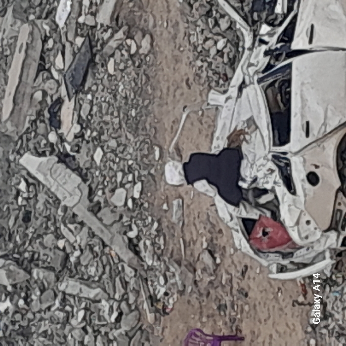 The image depicts a scene of extensive destruction in Khan Yunis, Palestinian Territory. A person, wearing a white head covering, a light-colored garment, and a dark vest, sits with their back to the camera on the severely damaged remains of a white car. The car's rear section is crushed, exposing a reddish internal component, and is partially covered by white material. To the left, a purple plastic chair stands on the sandy ground. The background is dominated by a dense field of gray concrete and stone rubble, indicative of widespread structural collapse.