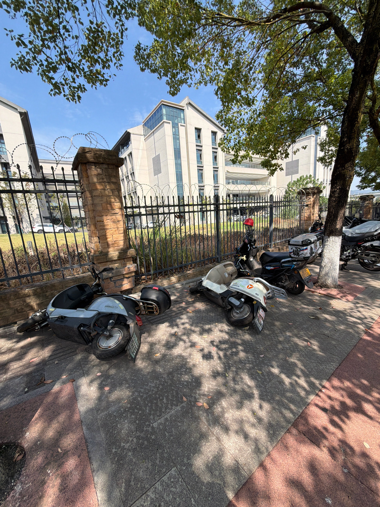 In Jiangjin, China, a bright, sunny day with a clear blue sky is depicted. The scene is set in an urban environment, likely a sidewalk or park area adjacent to a modern building complex. A black metal fence with barbed wire topping runs along the perimeter, with a brick pillar supporting a section of it.
Several parked vehicles, specifically scooters and motorcycles, are visible. One light-colored scooter is lying on its side on the sidewalk, near the fence. Another scooter, this one darker, is parked upright further down the path. Behind it, a black motorcycle is also parked. In the background, a person wearing a red helmet is standing next to a scooter, appearing to be stationary. The building in the background features white walls and glass-paneled windows. Lush green trees with shadows cast on the ground suggest the presence of foliage. The ground is paved with a mix of gray and reddish-brown tiles. Fallen leaves are scattered across the pavement.