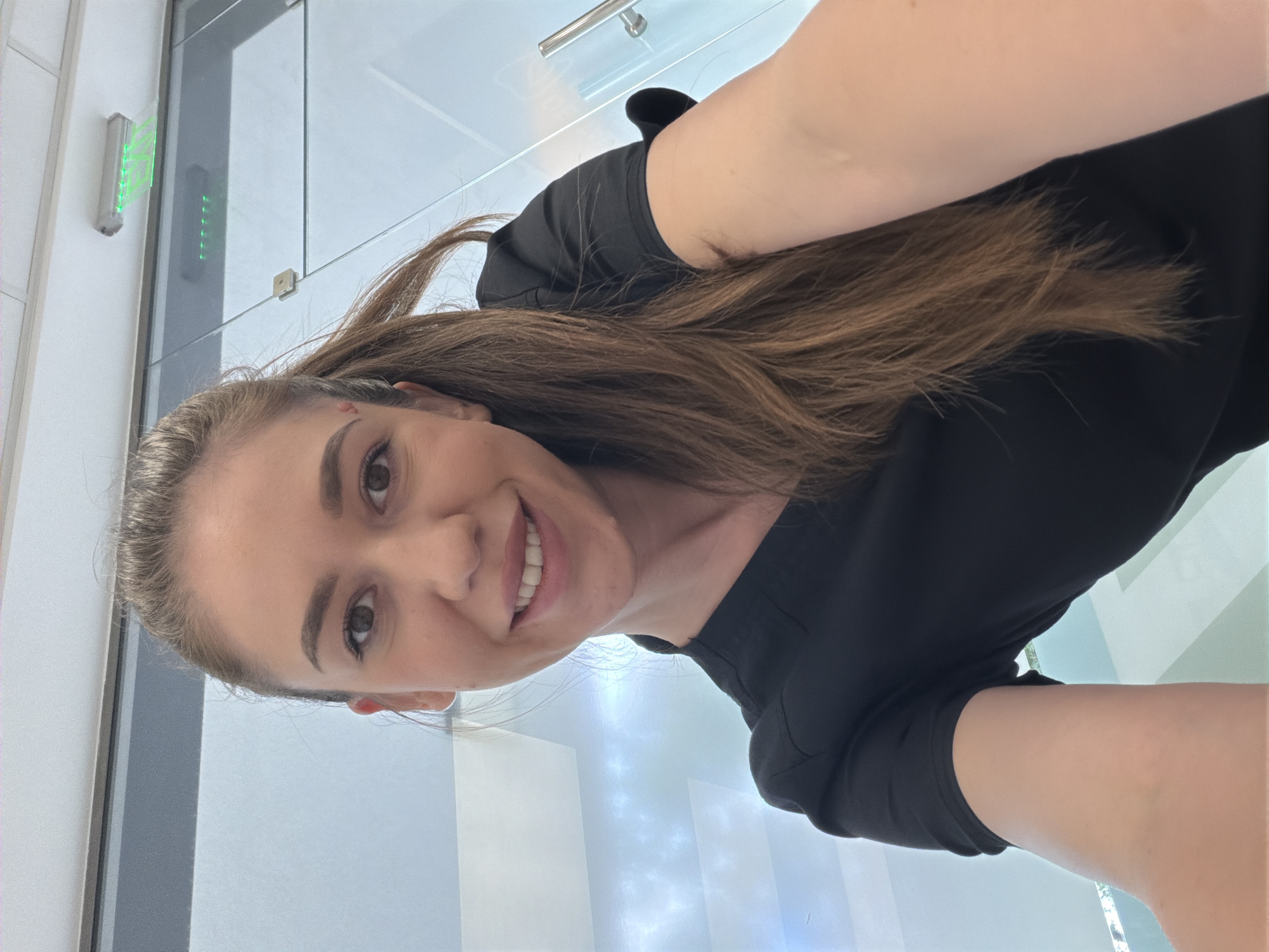 A young woman with long brown hair pulled into a ponytail smiles at the camera. She is wearing a black scrub top. A small red mark is visible on her left temple. She is in a brightly lit room with glass walls and ceilings that reflect a sky-like pattern. The background also shows what appears to be a ceiling with a light fixture. The overall impression is of an indoor, modern setting, possibly a clinic or office space, located in Sofia, Bulgaria.