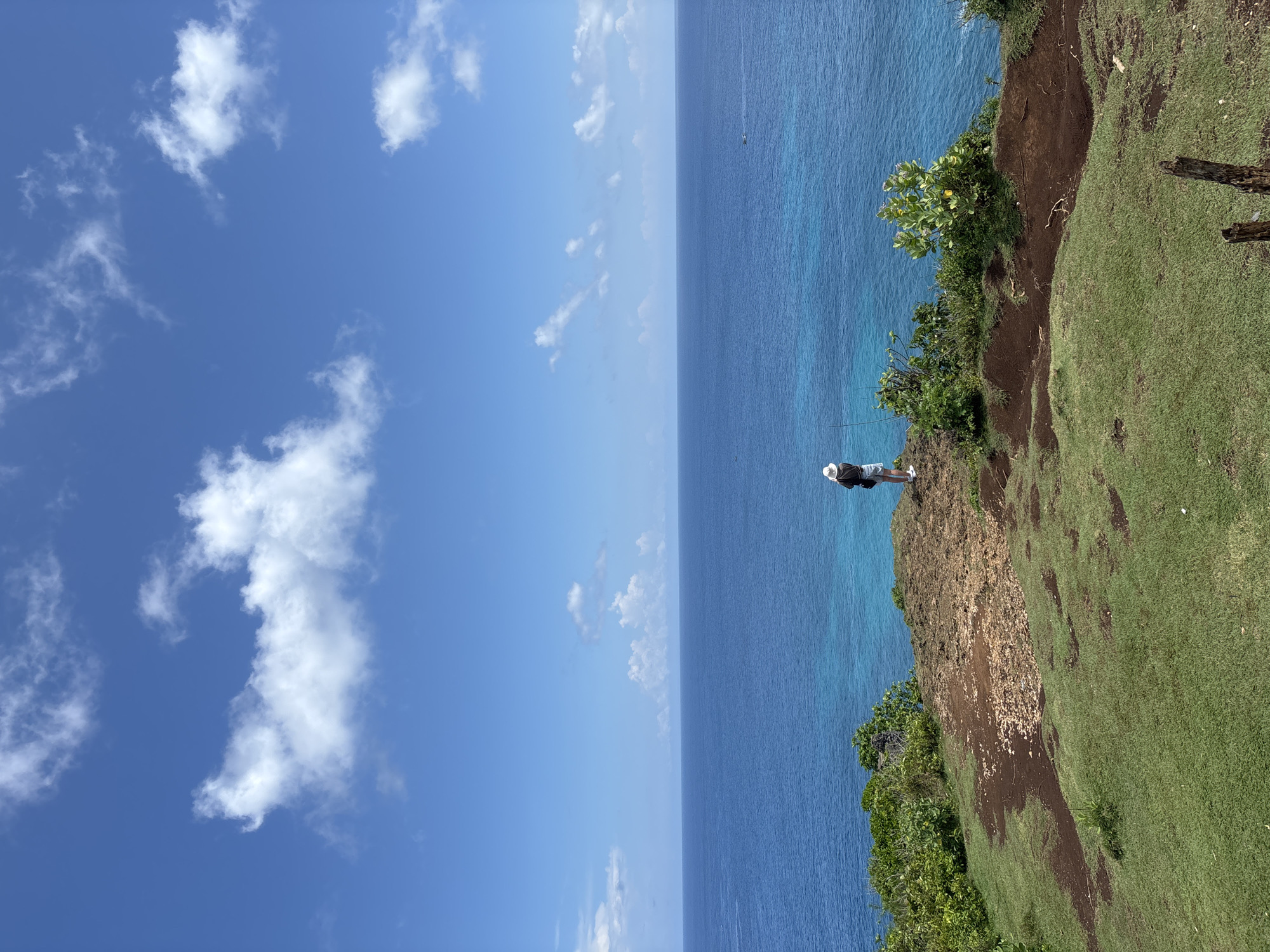 A vertical outdoor photograph captures a view in Uluwatu, Indonesia. A person stands on the edge of a grassy cliff overlooking the ocean. They are wearing a hat, dark jacket, and light shorts. The ocean extends to the horizon, meeting a sky with scattered clouds. Vegetation grows along the cliff's edge. Patches of bare dirt are mixed into the green grass, indicating erosion.