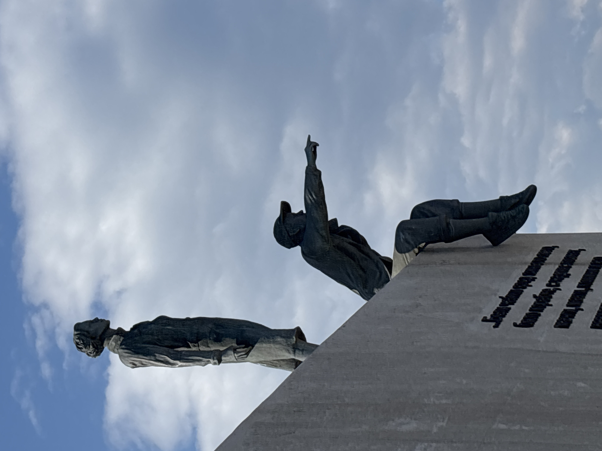 The image depicts a low-angle view of the Bulgarian-Soviet Friendship Monument in Varna, Bulgaria. The monument features two bronze statues of figures lying on a concrete structure. One figure points upwards. The background consists of a cloudy blue sky. There is text on a section of the concrete structure.