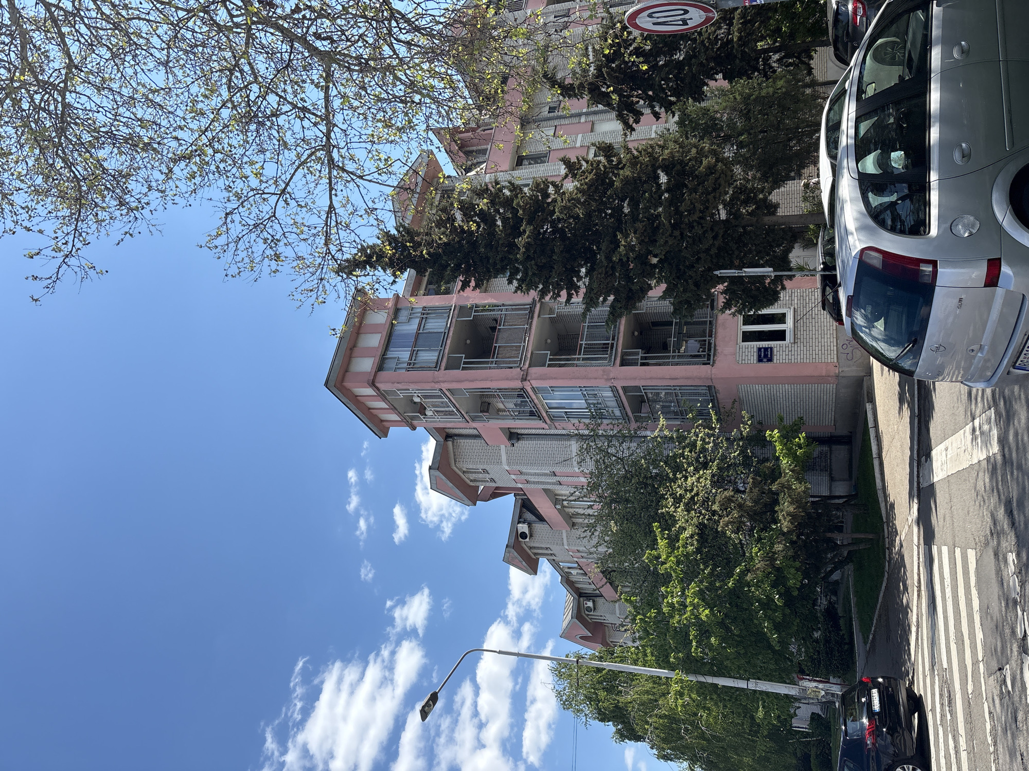 The image depicts a street scene in Belgrade, Serbia, featuring a light pink and white apartment building with balconies and multiple floors. A silver Opel Zafira passenger car is parked on the right side of the frame, partially obstructing the view of the building. A speed limit sign (40 km/h) is visible near the top of the building. Trees with green foliage and bare branches surround the building. The sky is bright blue with scattered white clouds. A street light is visible in the lower portion of the frame. A dark-colored passenger car is parked further down the street. A zebra crossing is visible on the street.
