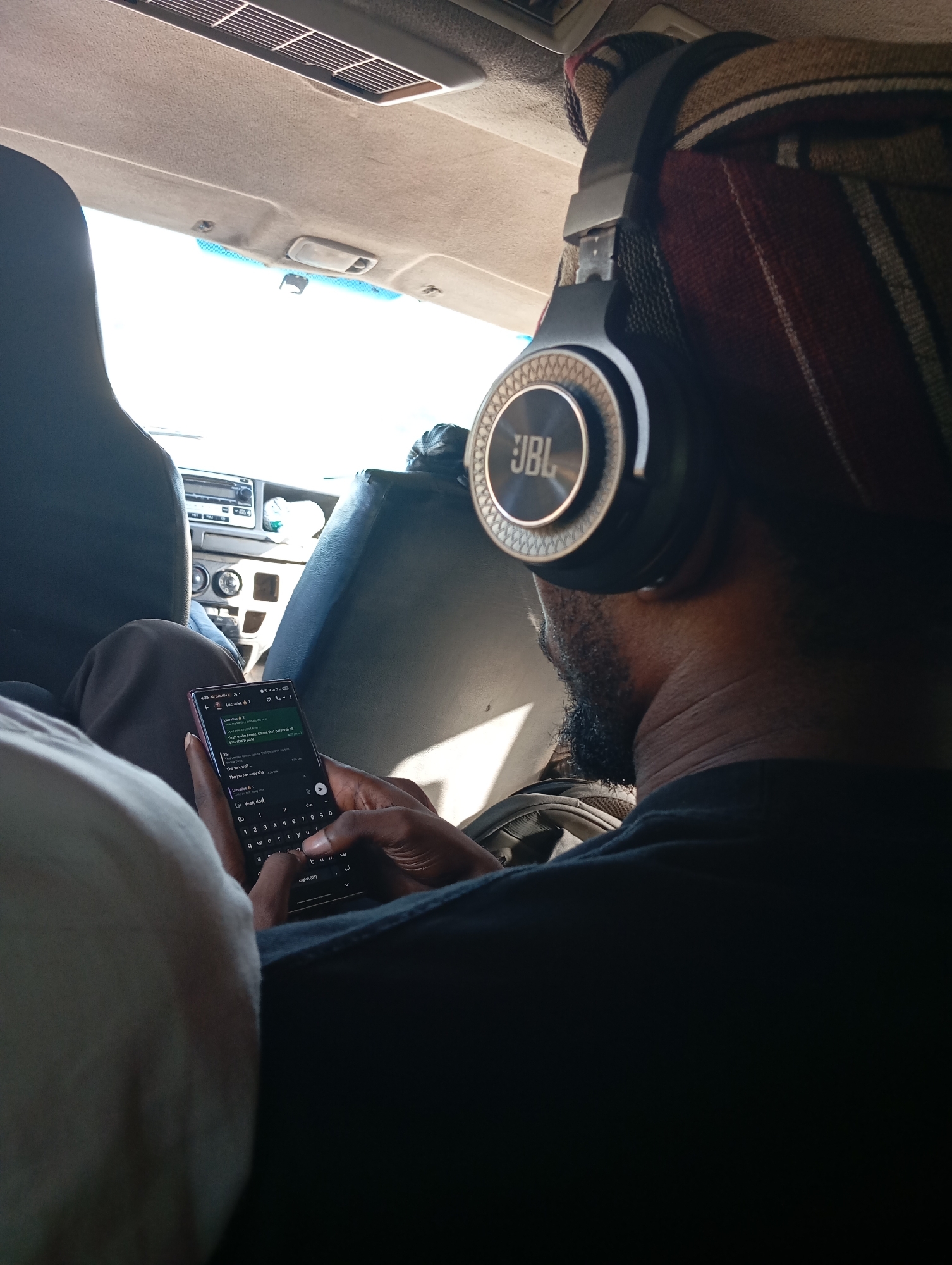 The image depicts a person with dark skin and a beard, wearing JBL headphones and a patterned head covering, seated inside a vehicle. The individual is looking at and typing on a smartphone, engaged in a text conversation. The car interior is visible, including the dashboard, seats, and ceiling features. The location implied by the smartphone screen is Gudi, Nigeria.