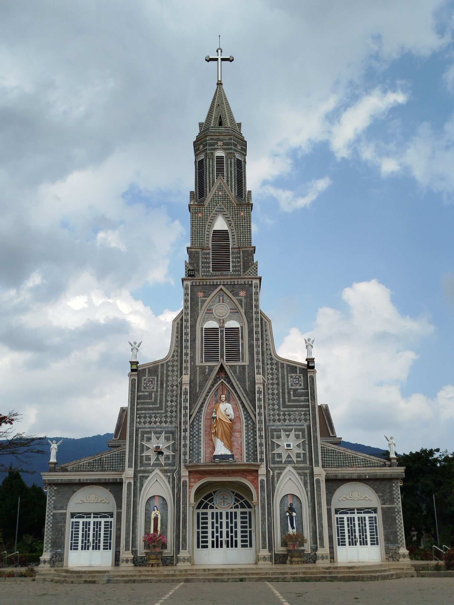 The image displays a prominent, ornate church building captured from a slightly low-angle, head-on perspective. The central subject is a grand stone church with a tall, multi-tiered steeple culminating in a cross at its apex. The church's facade is richly decorated with intricate geometric patterns in shades of grey, dark grey, and subtle off-white, giving it a patterned, almost mosaic-like appearance.
Above the main entrance, a large statue of a male figure, possibly Jesus or a saint, is prominently featured, clad in golden robes and holding a staff. Below this, flanking the white double doors of the main entrance, are two smaller statues recessed in arched niches: one appears to be a figure in blue and white robes, possibly the Virgin Mary, while the other depicts a male saint in dark robes. Two white angelic figures with outstretched wings are positioned on the lower roof sections, one on each side of the central tower. The church features multiple arched windows with louvred vents and large white double doors with intricate paneling.
In the background, lush, dark green trees frame the sides of the church, and distant mountains are visible under a partly cloudy sky. The sky transitions from a heavily clouded, light grey on the left to patches of clear blue with scattered white clouds on the right. The foreground shows a paved area in front of the church, with steps leading up to the entrance.
The scene takes place during daylight hours, under fair but partly cloudy weather conditions. No specific activity or event is captured, and no discernible text is visible on the church or in the surrounding environment. The image is set in Giãn Dân, Vietnam.