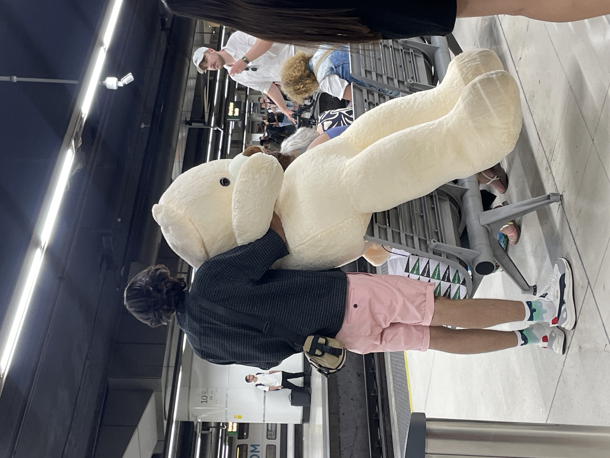 A person stands on a platform at a Barcelona metro station, holding a very large, cream-colored stuffed bear. The person is wearing a dark, checkered shirt, pink shorts, and athletic shoes with colorful socks. A tan shoulder bag is slung across their body. The bear is positioned in front of them, its legs extending down towards the floor. Other people are visible in the background, some sitting on benches and others standing. A train is visible on the tracks to the left. The environment appears to be indoors with overhead lighting and a dark ceiling.