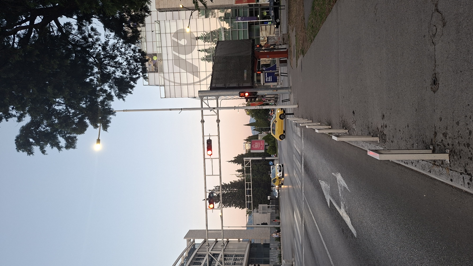 The image shows a street scene in Podgorica, Montenegro, near the intersection of Stanka Radonjića Street and Bulevar Džemala Bijedića. A traffic light is red for eastbound traffic. Two yellow cars are visible, one a hatchback and the other a sedan. A building with a large "70" logo on its facade is visible in the background. The sky is clear and the sun is setting, casting a warm glow on the scene.