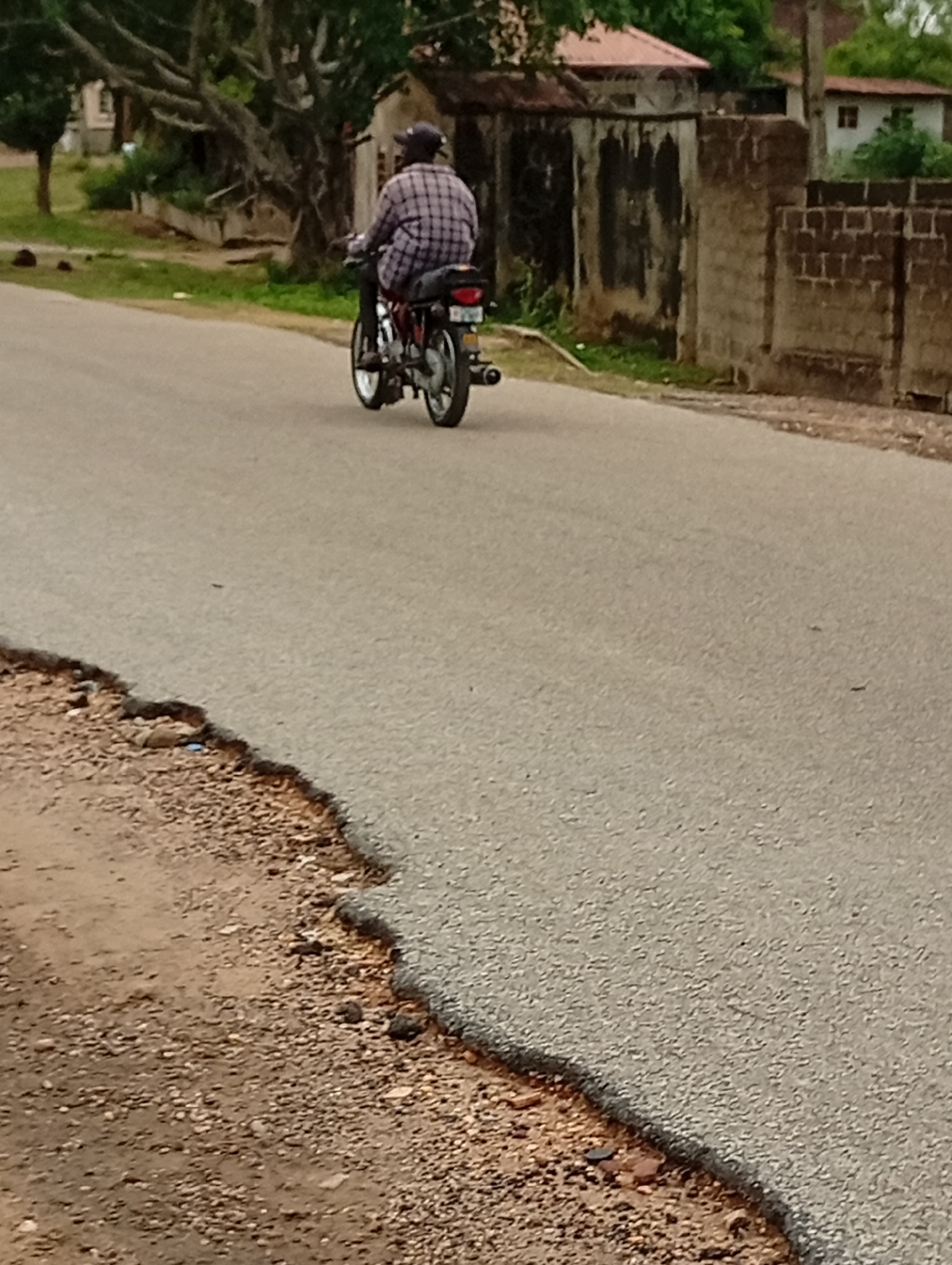 A man rides a motorcycle on a paved road in Lokoja, Nigeria. He wears a plaid shirt and a cap. The road has damaged edges exposing gravel. Buildings and vegetation line the roadside.