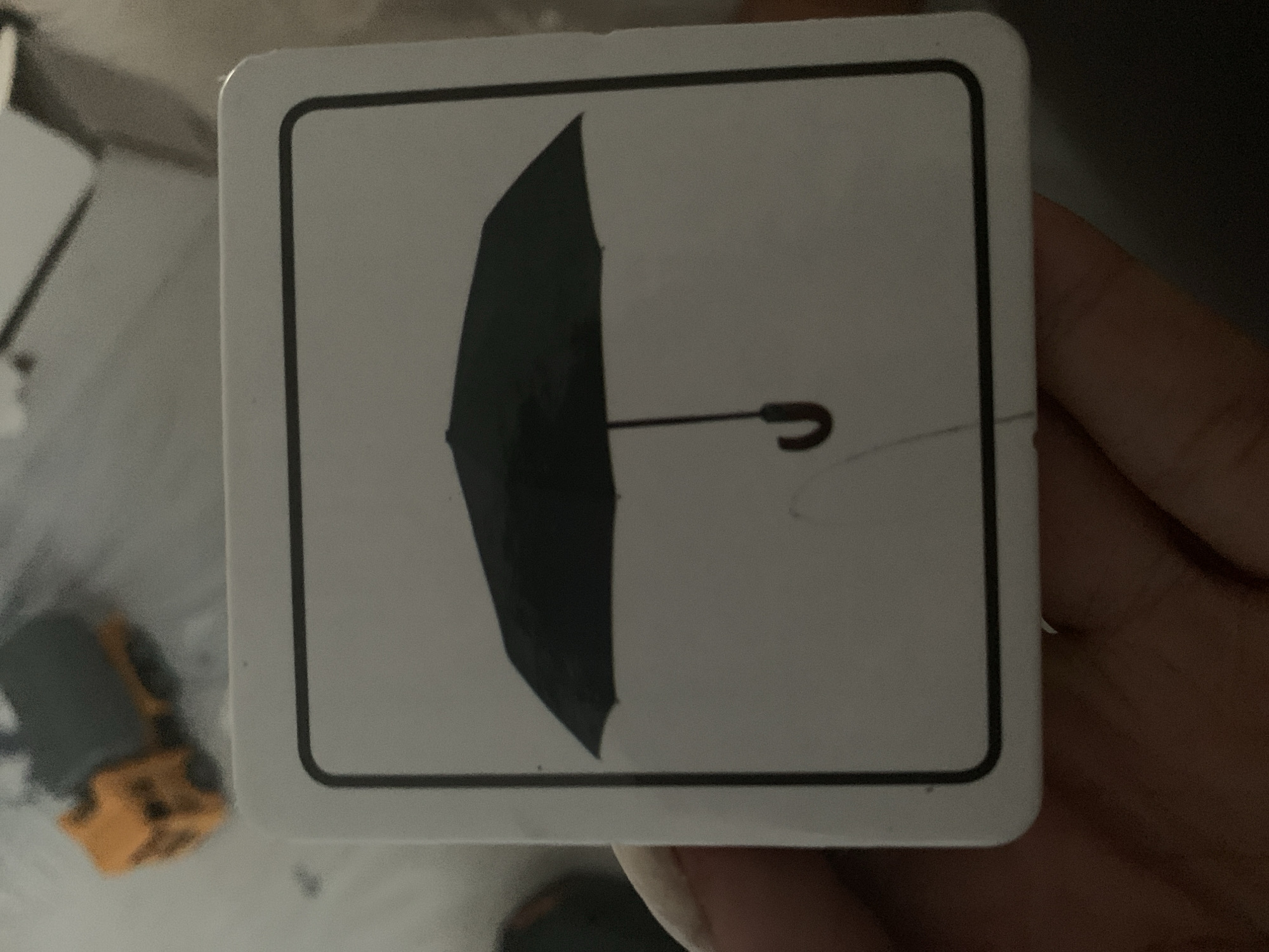 The media file is a close-up, angled shot of a white card with a black outline. Inside the outline is a silhouette of a closed black umbrella. The umbrella is oriented vertically with the handle on the right side, curving upwards. The card is held by a person's hand, visible on the right side of the frame. The background is out of focus, but appears to be an indoor setting with some blurry objects, including what looks like a yellow toy in the lower left corner. The lighting is dim, casting shadows and suggesting it might be indoors or evening. There is no visible text on the card.