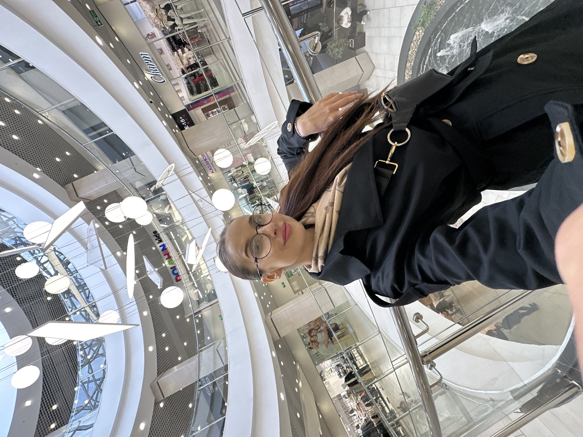 The image, oriented 90 degrees counter-clockwise, depicts a woman taking a selfie inside a brightly lit, multi-story shopping mall in Yekaterinburg, Russia. The woman, in the foreground, has long brown hair, wears glasses, a black coat with gold buttons, and a patterned scarf. A black bag with prominent gold hardware hangs from her shoulder. Her expression is neutral, showing a slight smile.
The setting is a spacious, modern atrium characterized by soaring curved white architectural elements, extensive glass railings, and reflective surfaces. The space is illuminated by numerous large, circular and rectangular overhead lights. In the lower right, a dark-colored water feature or fountain is visible. Reflections of people and the building structure can be seen on the polished floor.
Numerous shop signs are visible on various levels. Identifiable text includes "Charm", an "E 18+" age restriction sign, "DOLPHIN kids", and "КАЗАНОВО" (Kazanovo). Other Cyrillic text visible on signs includes "МЕХА И КОЖА" (Furs and Leather), "МОДНАЯ ОДЕЖДА ИЗ ГЕРМАНИИ" (Fashionable Clothing from Germany), and an exit sign with an arrow. The scene conveys a bustling, contemporary indoor commercial environment, likely during daytime.