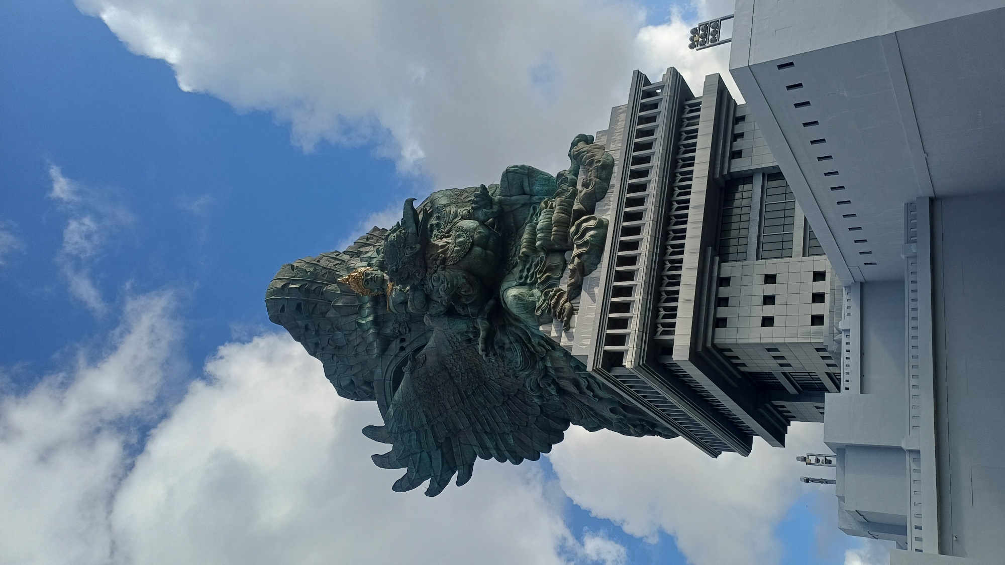 The image shows the Garuda Wisnu Kencana statue in Kuta Selatan, Indonesia. The statue, primarily composed of dark green bronze, depicts the Hindu god Vishnu riding the mythical bird Garuda. The statue's head, shoulders, torso, and wings are visible. It is situated beside a tall, rectangular building. The building has a grey facade with a grid of windows. Parts of a white building can be seen to the right of the grey building. The sky is visible in the background, with a mix of blue sky and white clouds.