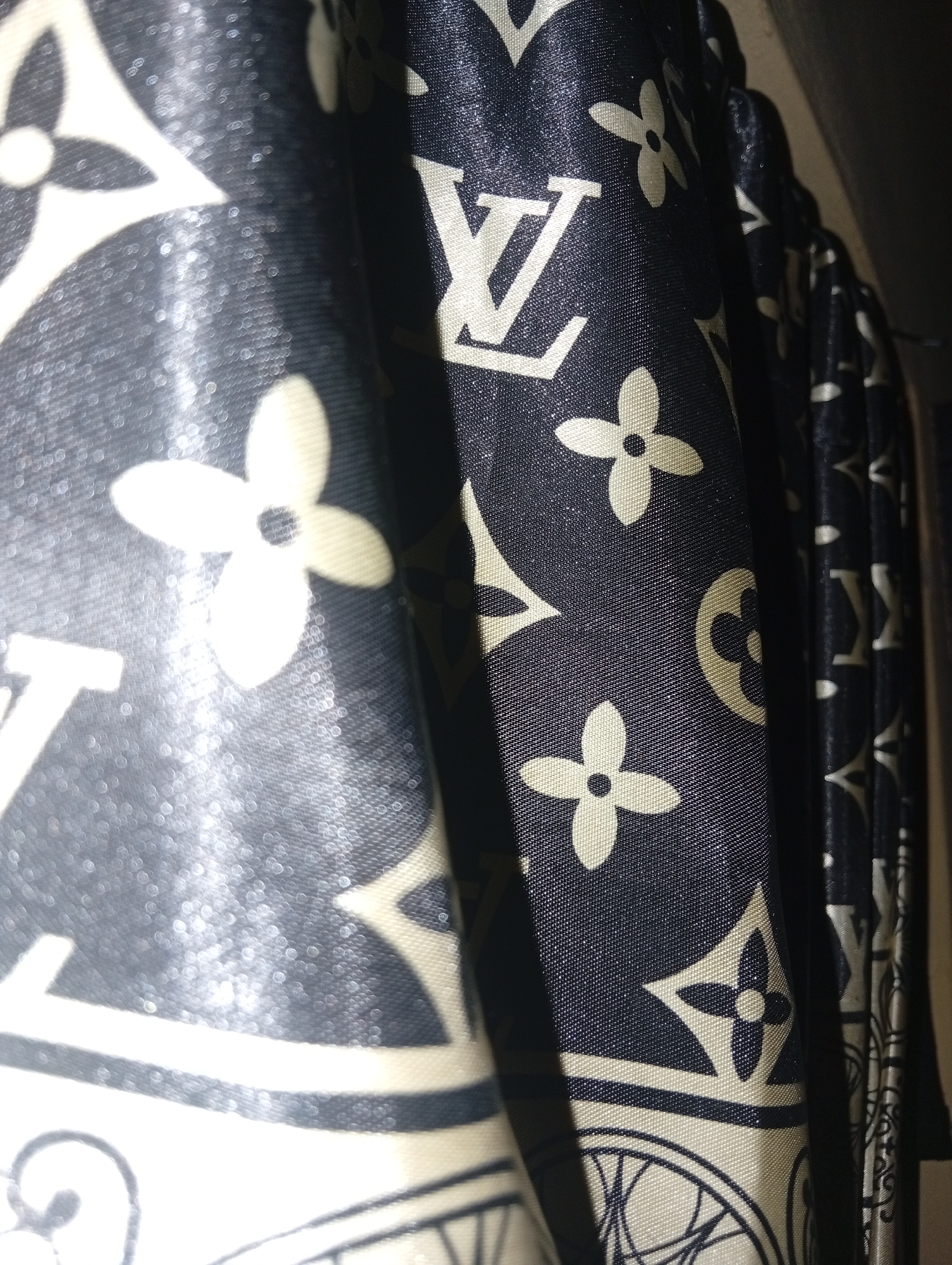 The image shows a close-up view of a dark blue fabric with a repeating pattern of beige Louis Vuitton monograms. The fabric appears to be curtains or a similar textile, with folds and creases creating variations in light and shadow. The monograms, including the LV logo and the four-petal flower, are clearly visible and have a slightly metallic sheen. The background is dark and out of focus. No people, actions, or interactions are depicted. The image does not provide any information about its location in Ukukwa, Nigeria.